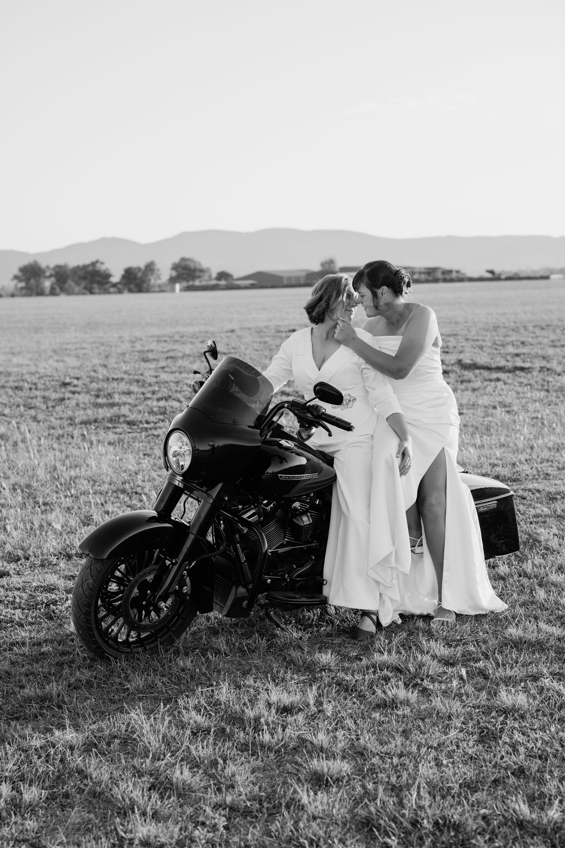 Two women in wedding dresses beside a black motorcycle in a field, looking at each other, mountains in the background.