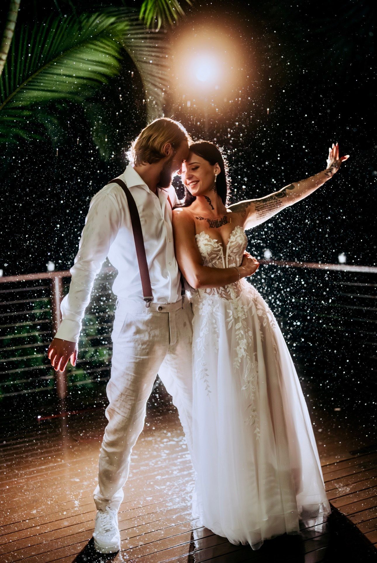 Couple in wedding attire dances outdoors under rain. Night setting with lights and foliage.