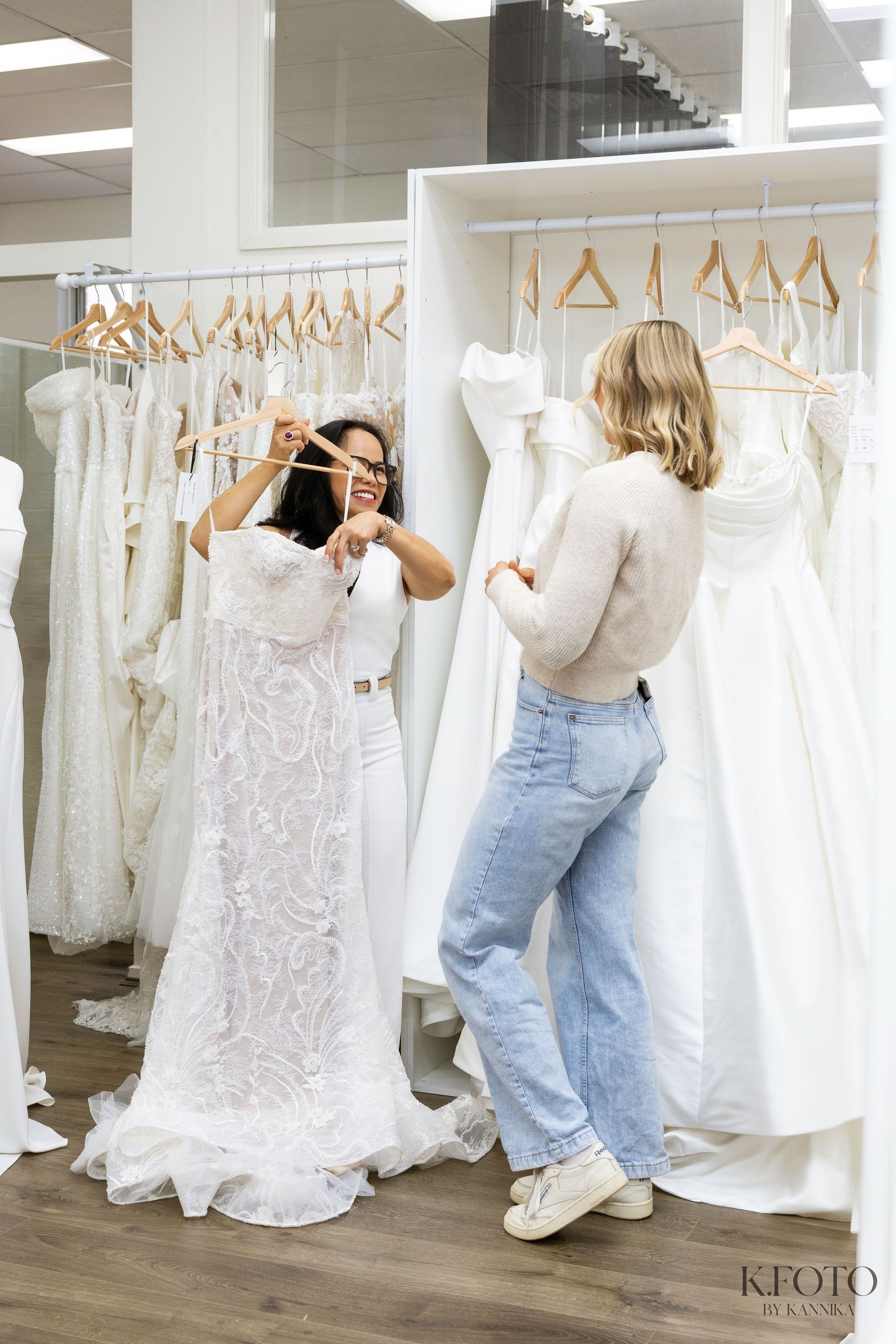 Two women are standing in front of a closet full of white dresses.