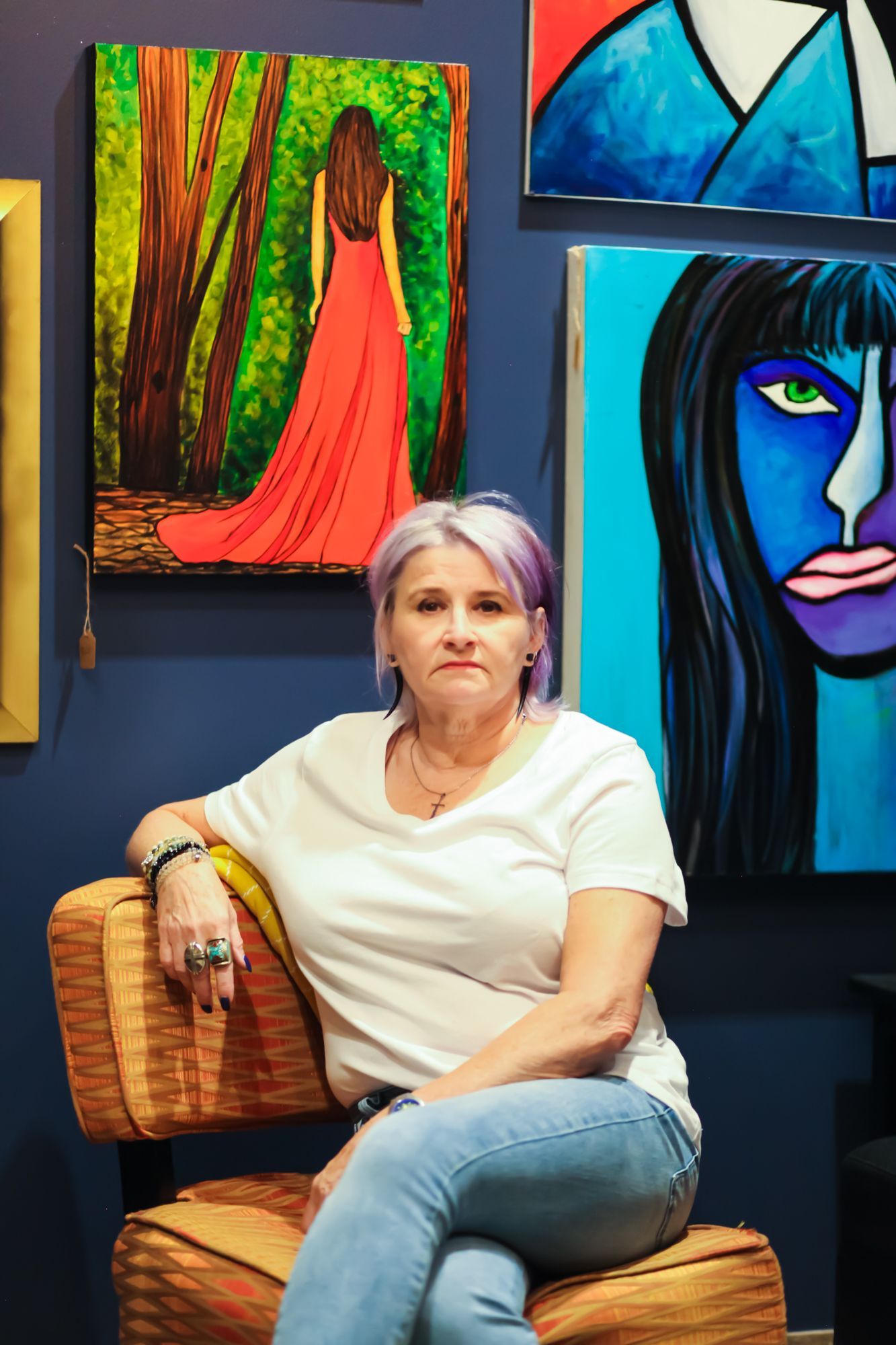 Woman seated in a chair, with purple-streaked hair, in front of colorful artwork on a blue wall.