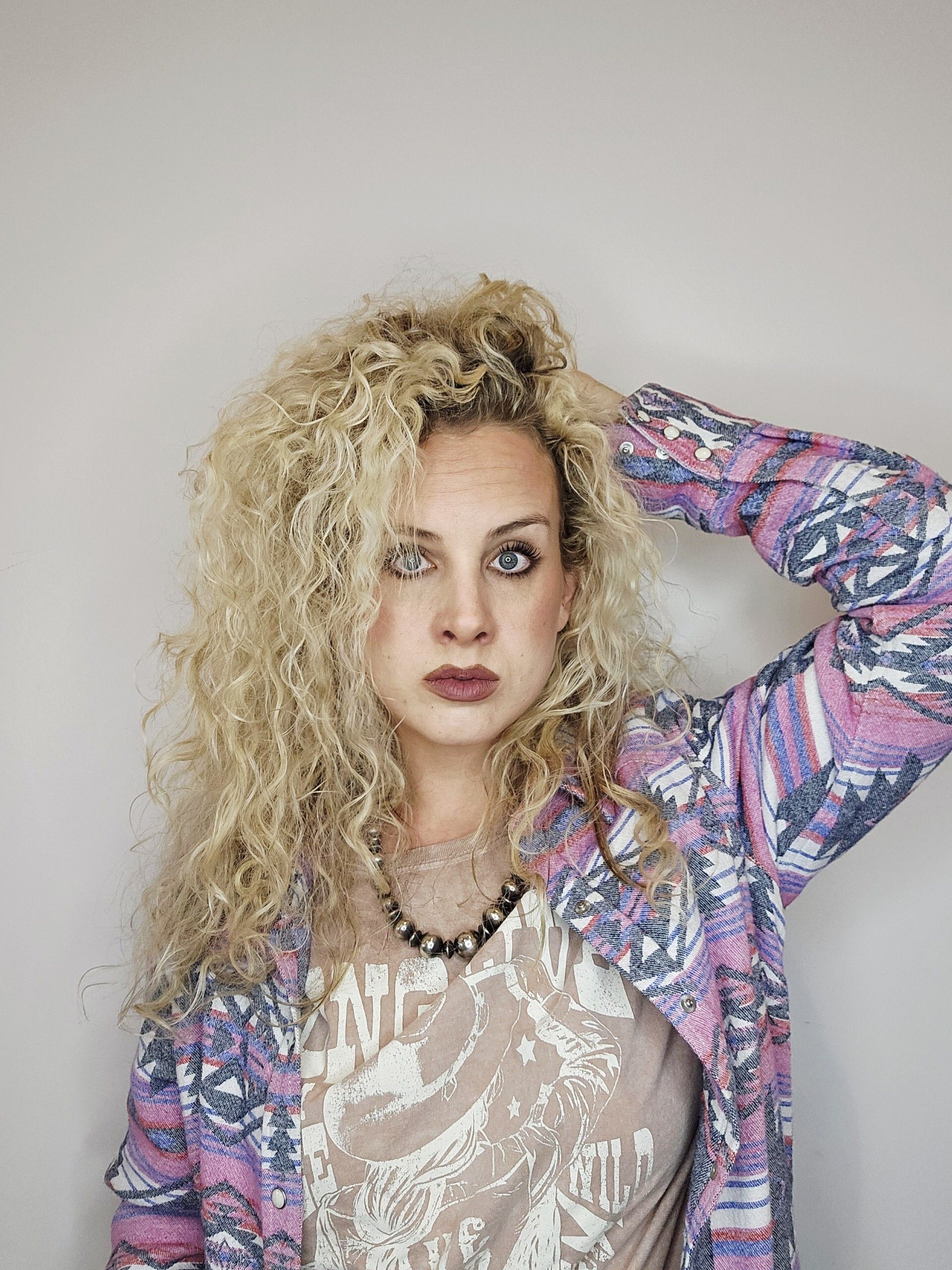 Woman with curly blonde hair, hand in hair, wearing patterned jacket and necklace, neutral expression.