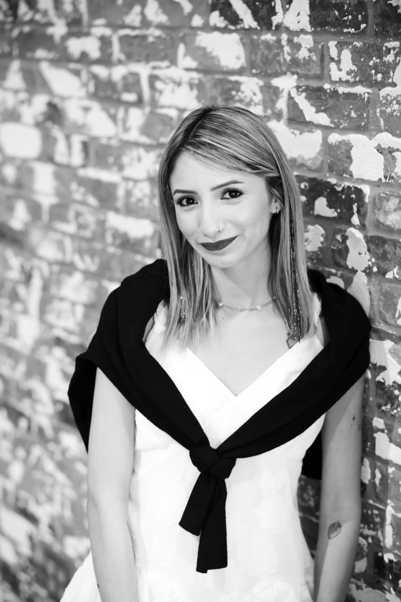 Woman wearing a white dress and black sweater, leaning against a brick wall.