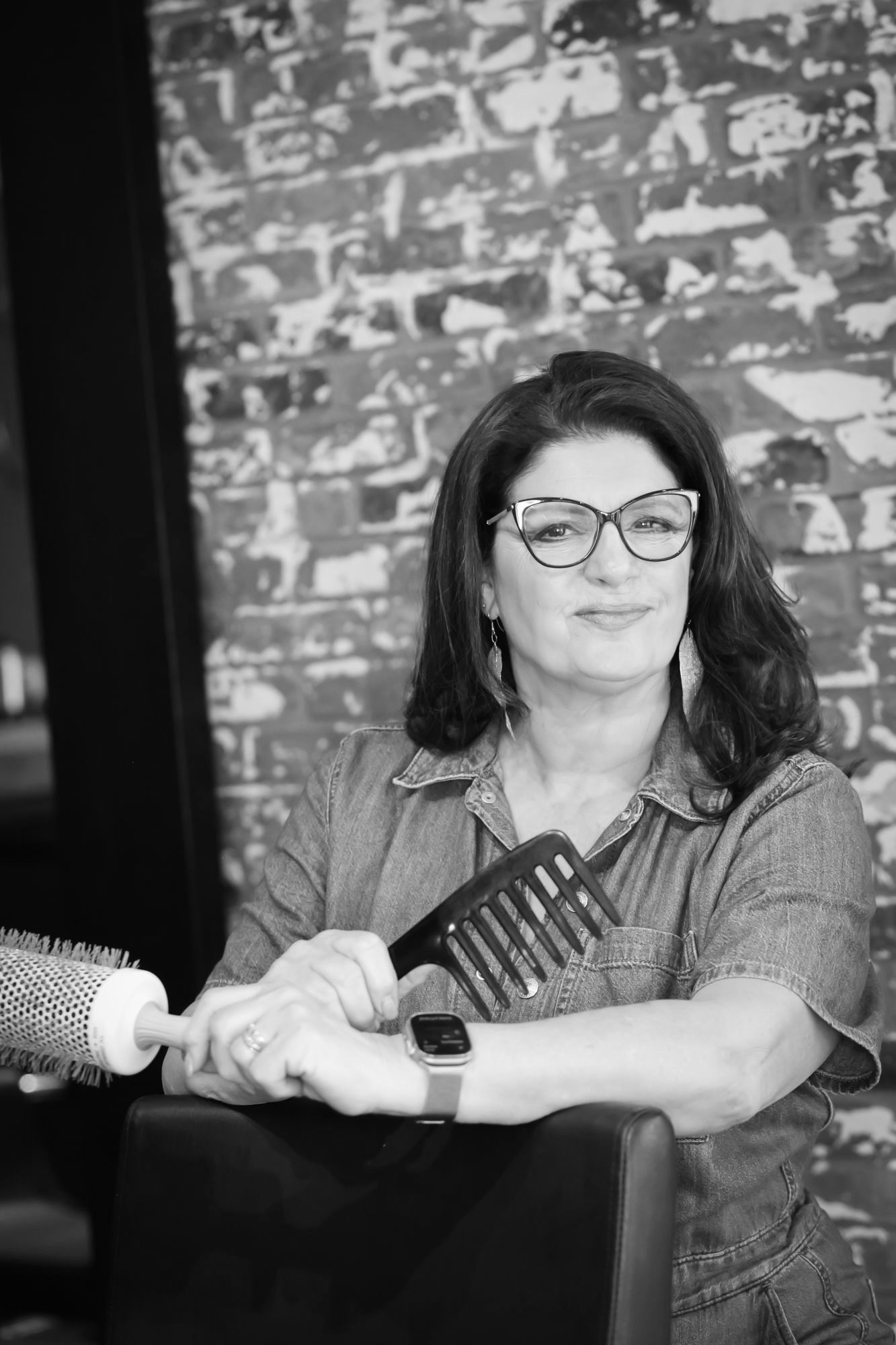 Woman with glasses holds a comb and brush, sitting in a salon chair. Brick wall background.