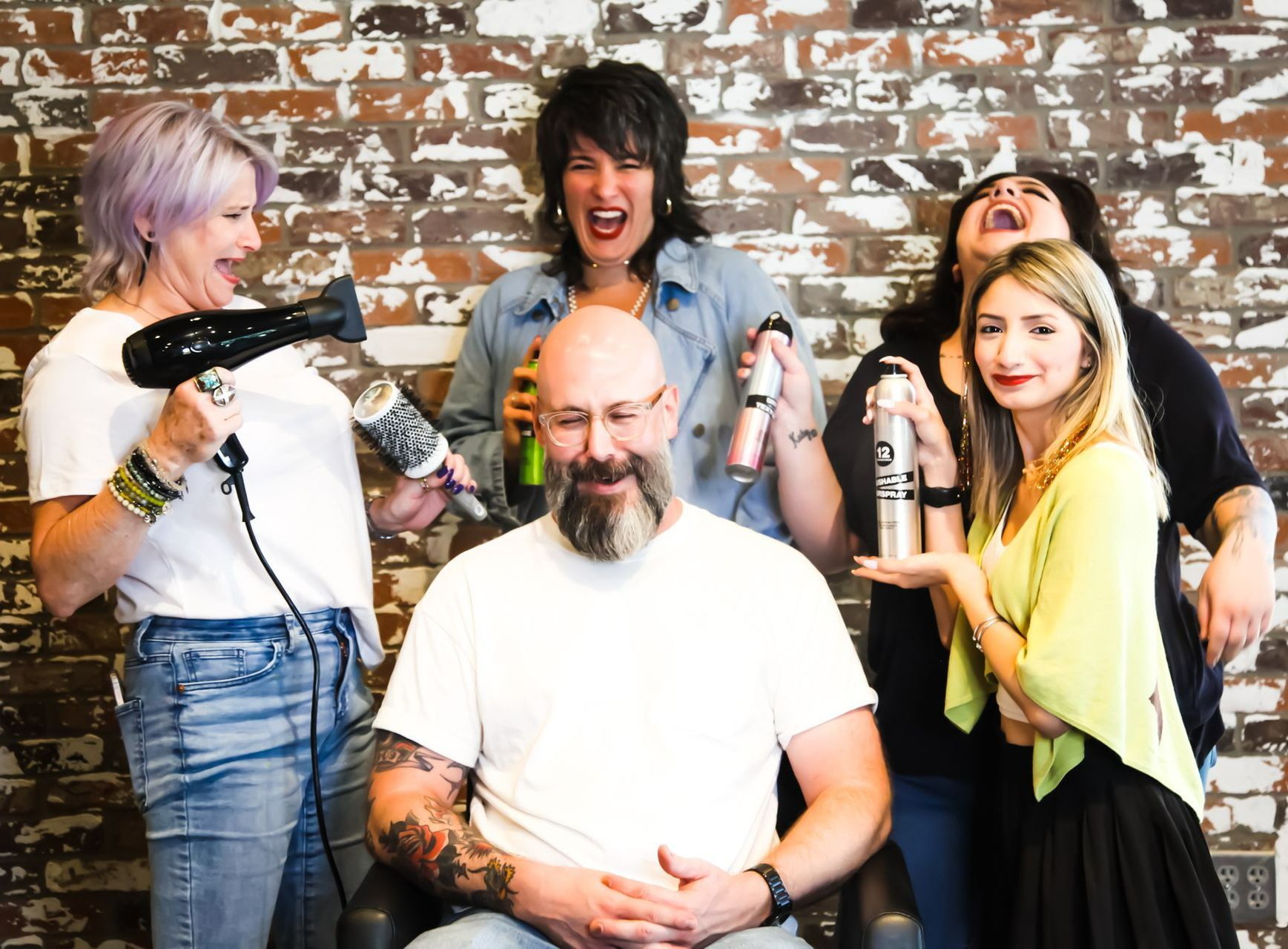 Hair stylists playfully surround a man in a chair, holding styling tools, laughing against a brick wall.