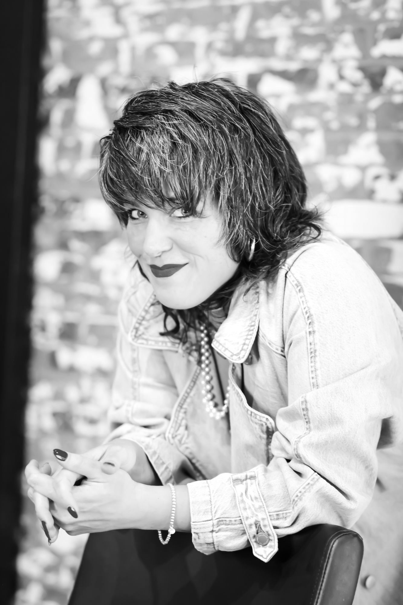 Woman leaning on a chair, wearing a denim jacket and necklace, smiling. Brick wall background.