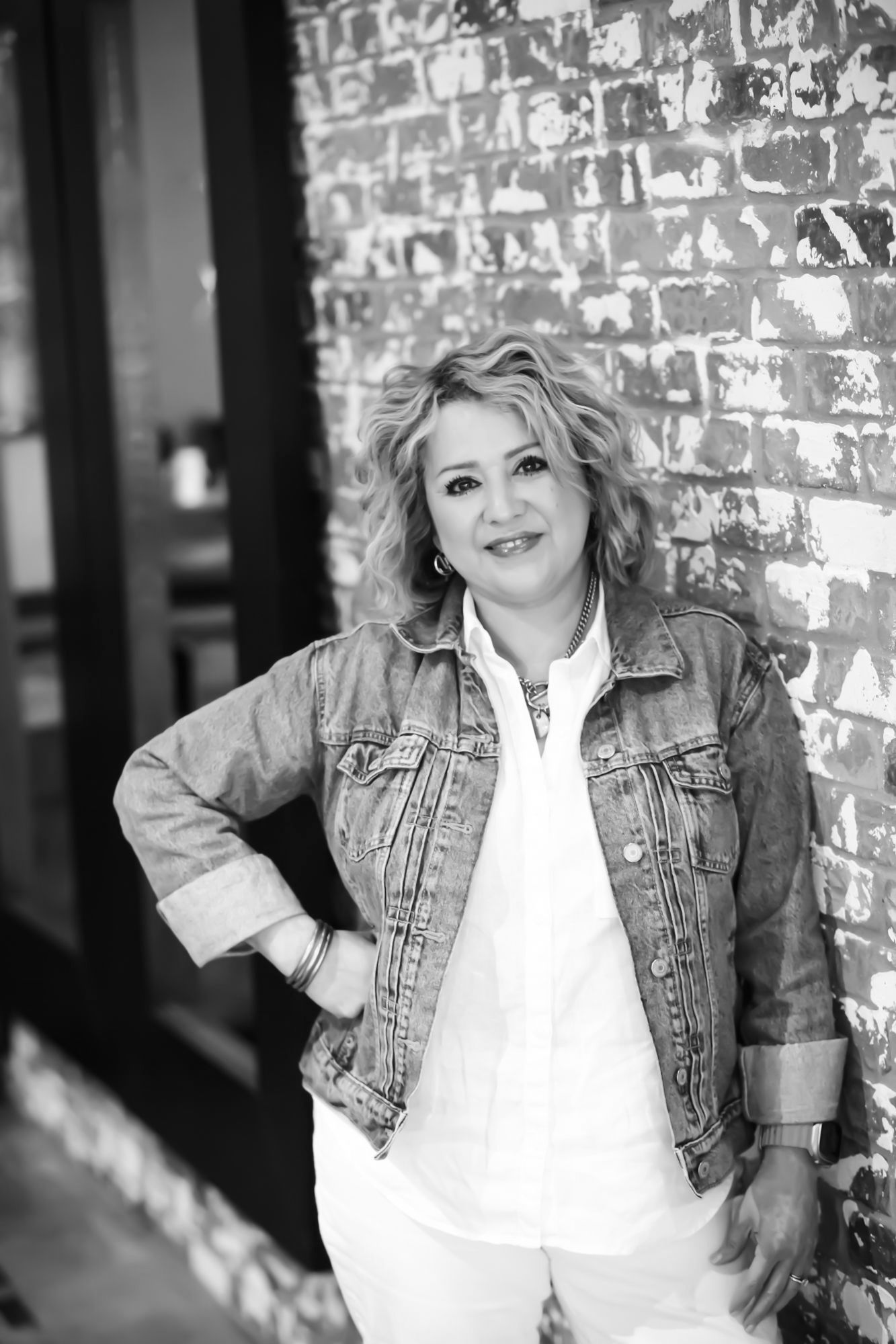 Woman leaning against a brick wall, wearing a denim jacket and white shirt, smiling.