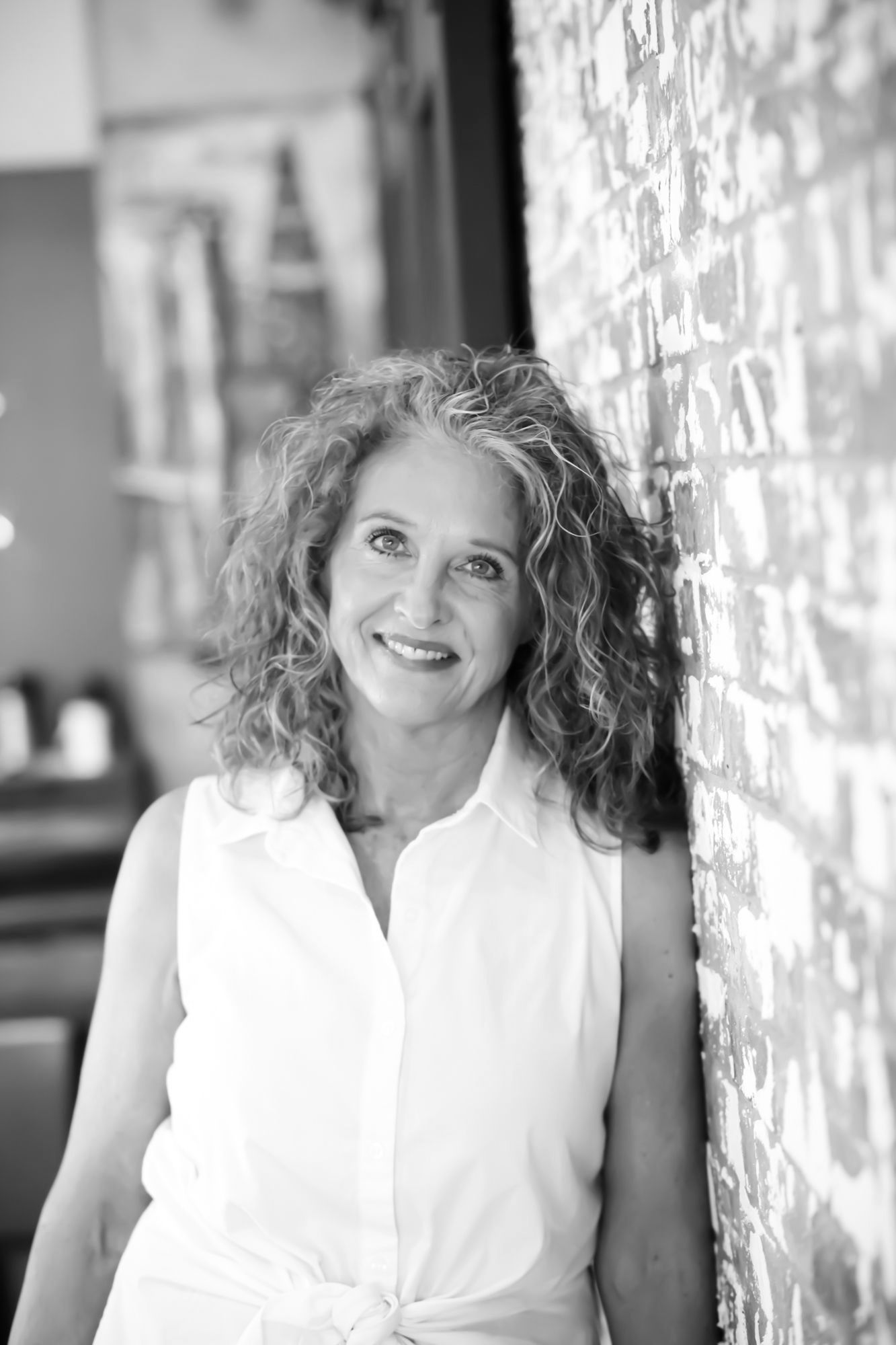 Woman with curly gray hair smiles while leaning against a brick wall, wearing a white sleeveless shirt.