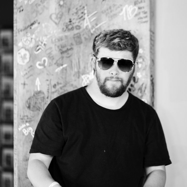Man in sunglasses and black shirt, standing in front of a graffiti-covered wall, looking towards the camera.