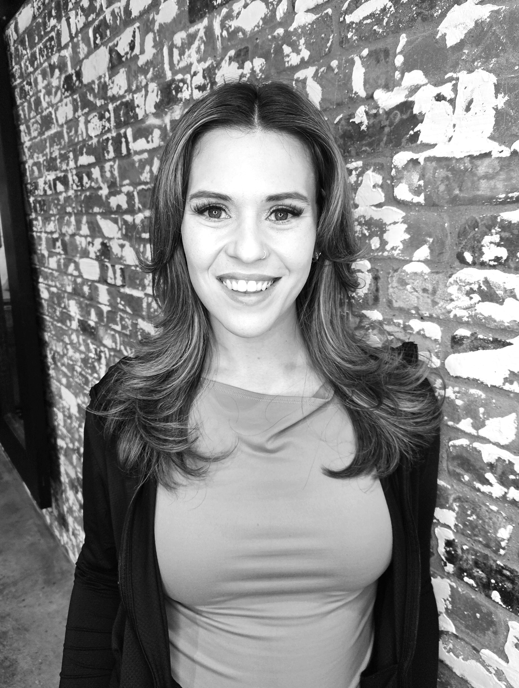A smiling person with long hair stands in front of a textured brick wall, wearing a top and an open jacket.