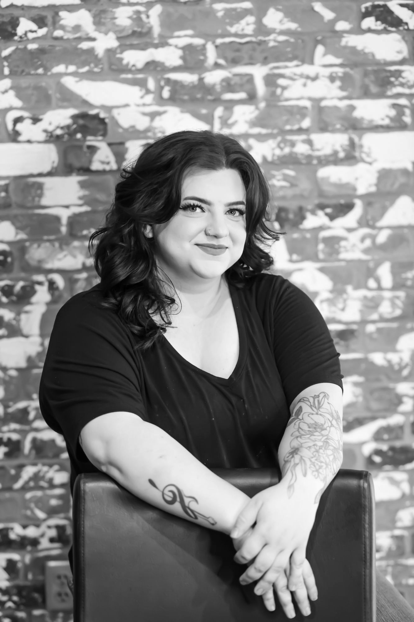 Woman with arm tattoos, smiling, sitting in front of a brick wall.