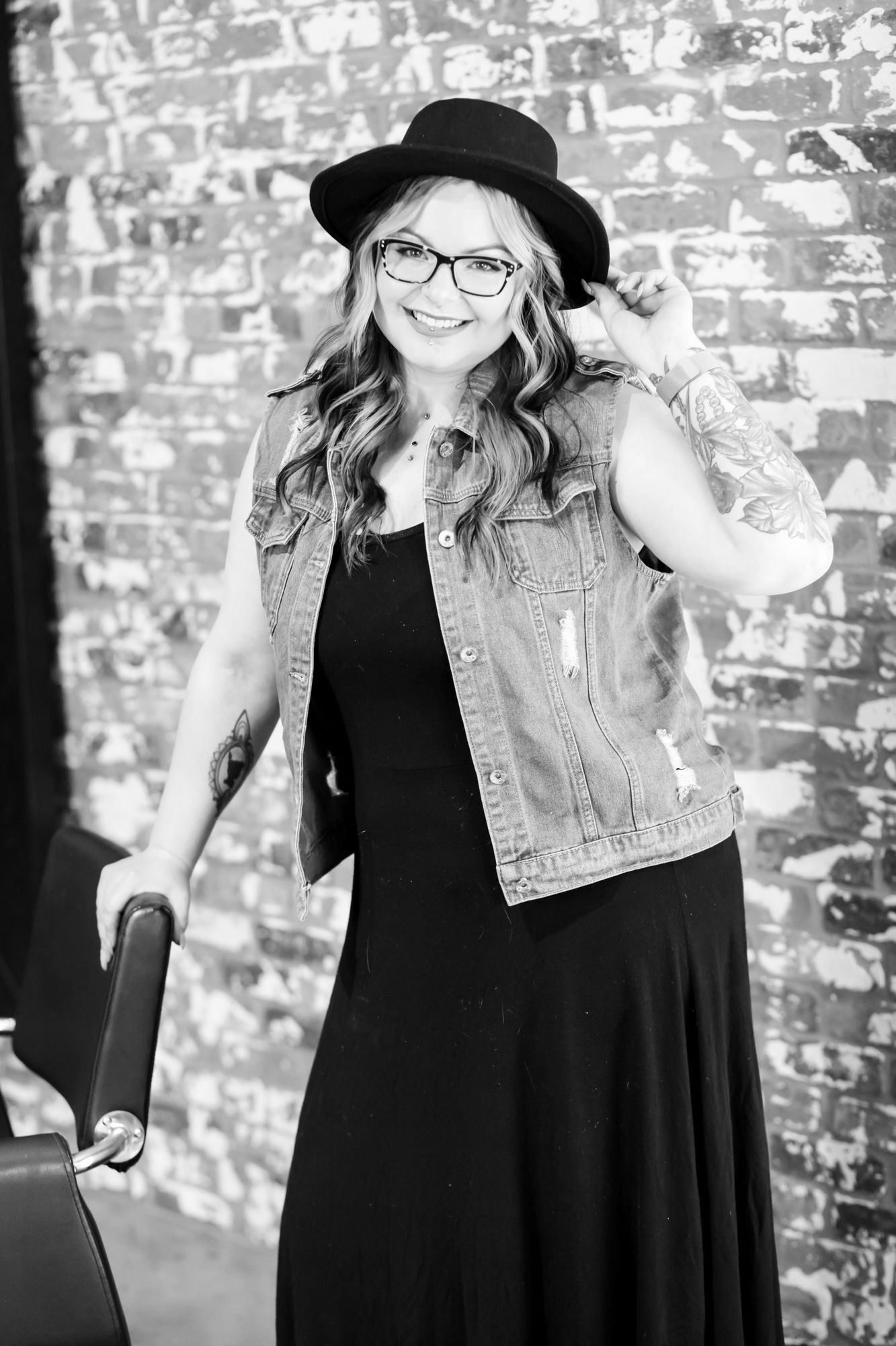 Woman wearing a hat, glasses, and denim vest, leaning on a chair in front of a brick wall.