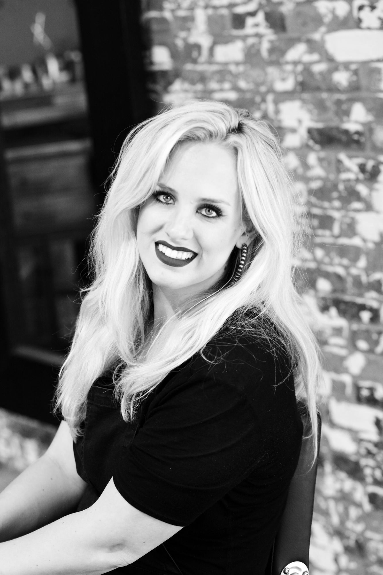 Blond woman smiles, dark lipstick, black shirt. Leans toward camera, brick background.
