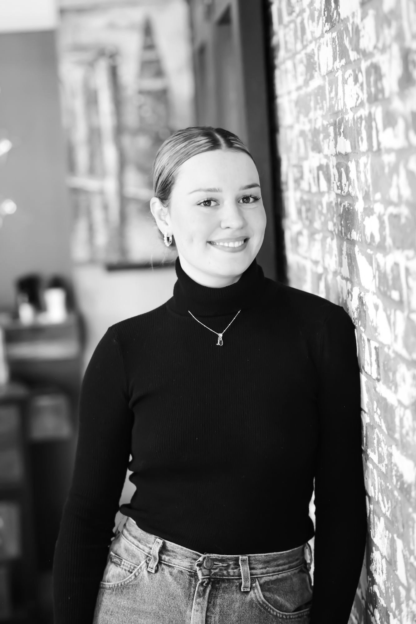 Woman in black turtleneck smiles, leans against brick wall.