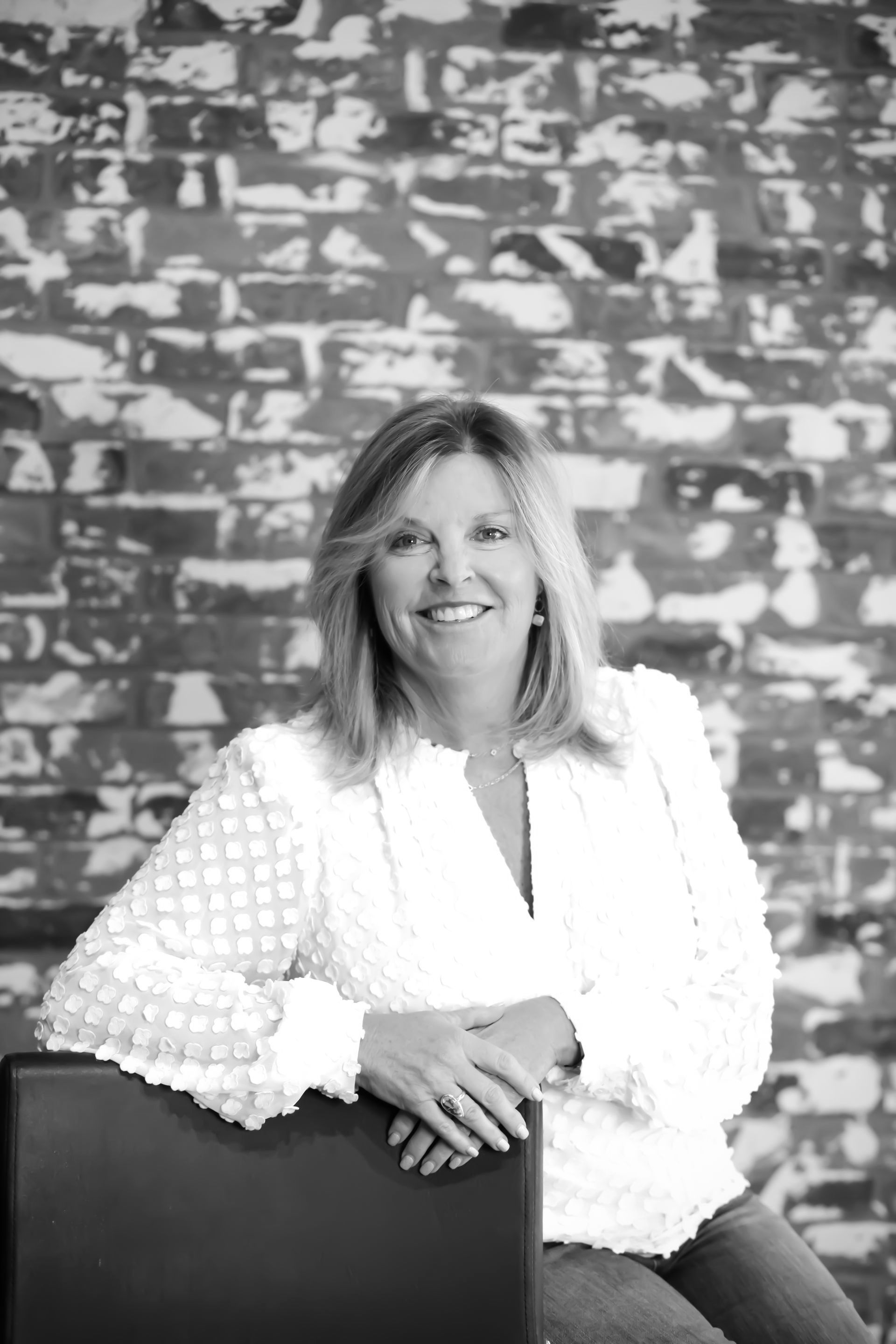 Woman in white shirt and jeans smiles, leaning on a chair in front of a brick wall.