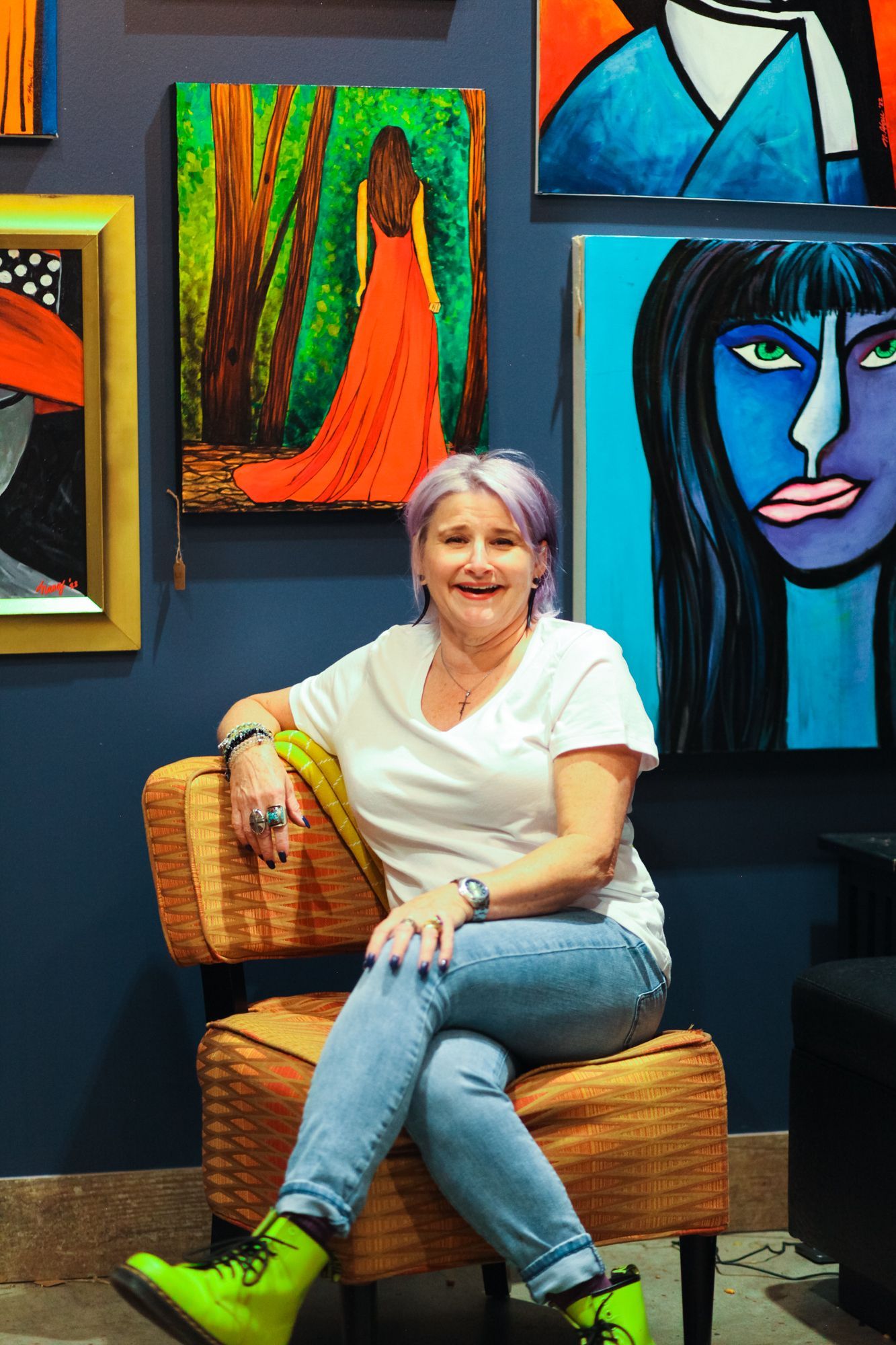 Woman with purple hair sits in a chair, smiling, surrounded by colorful abstract paintings.