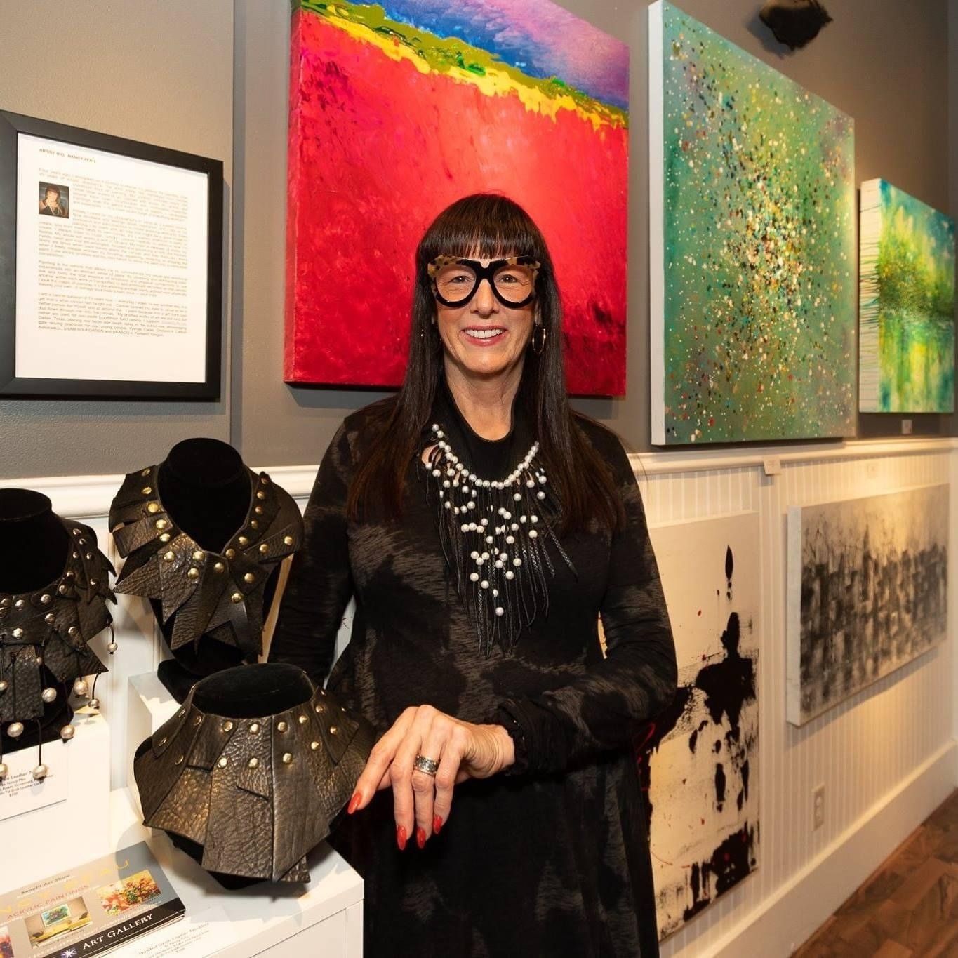 Woman in gallery with art and jewelry. Red, green, and black abstract paintings on wall behind her.
