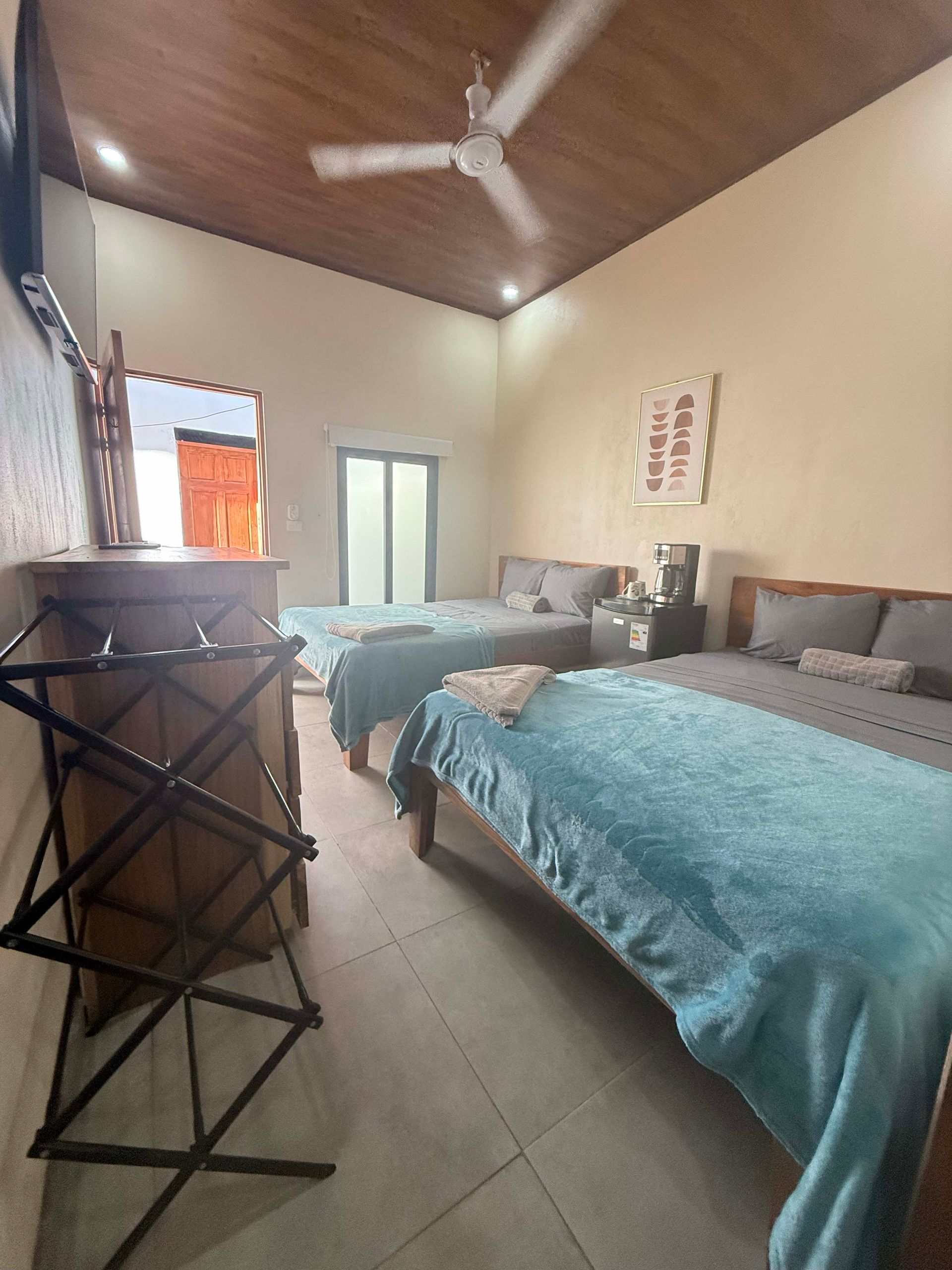 Hotel room with three beds, a luggage rack, and ceiling fan. Light blue bedspreads, wooden furniture.