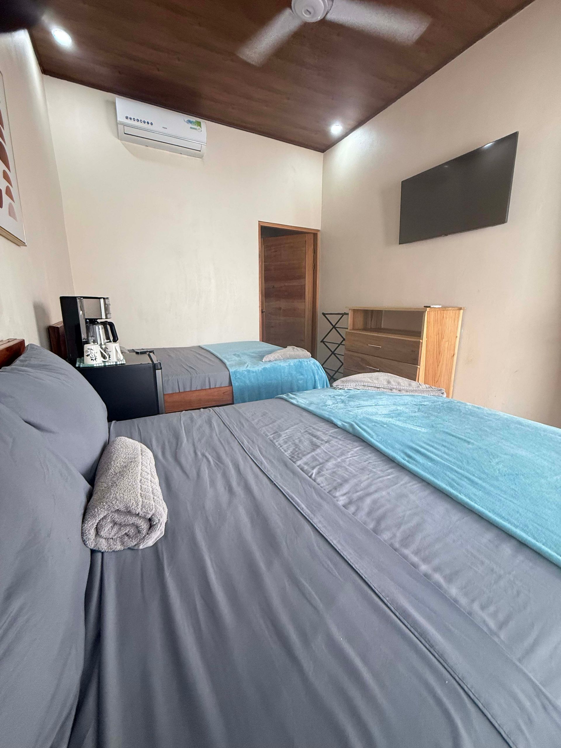 Bedroom with two beds, gray sheets, blue blankets, and wooden ceiling.
