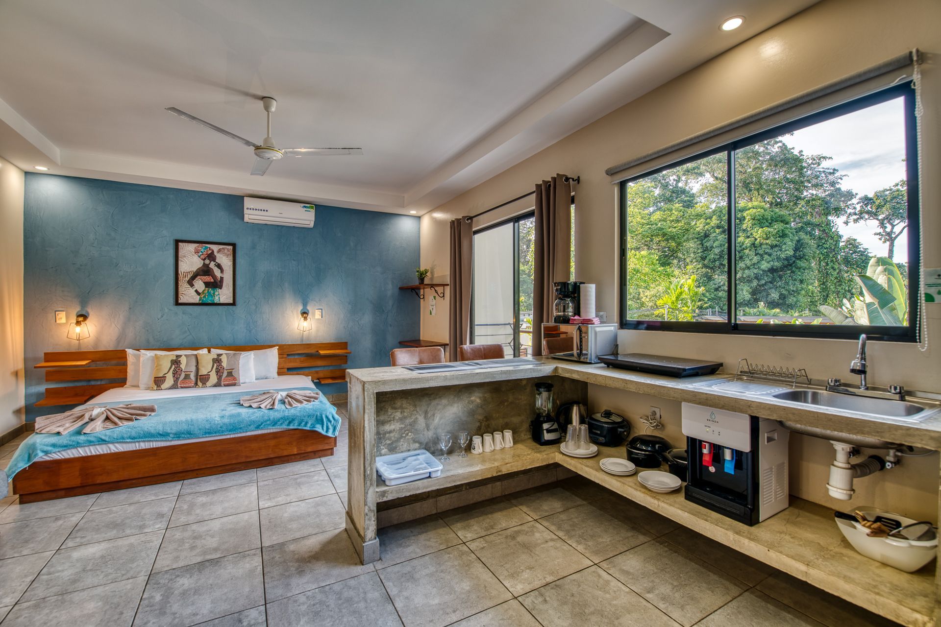 Bedroom with a bed, kitchen, and balcony with open doors overlooking greenery.