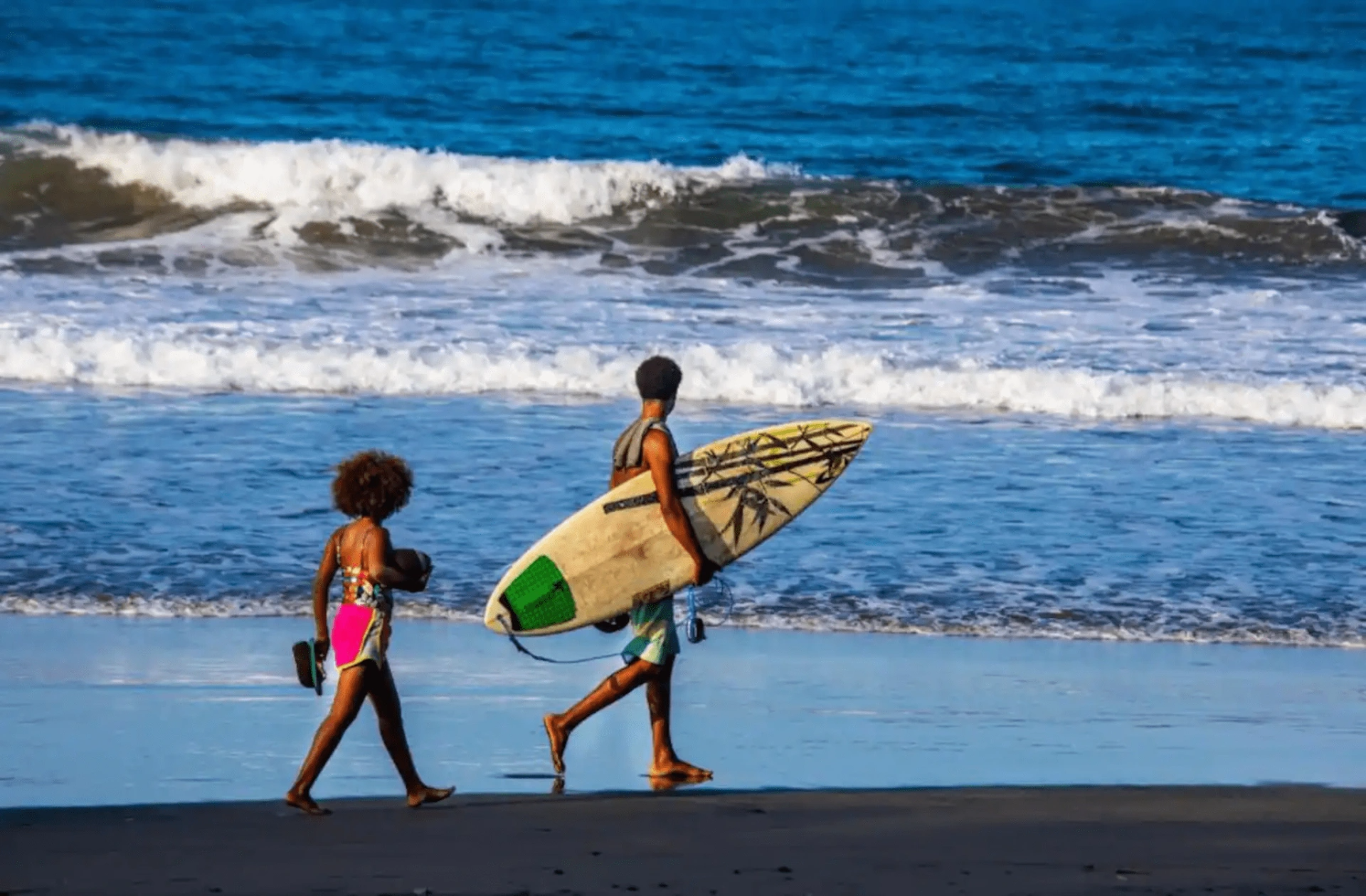 Two people walking on beach; one carrying a surfboard, waves in background.