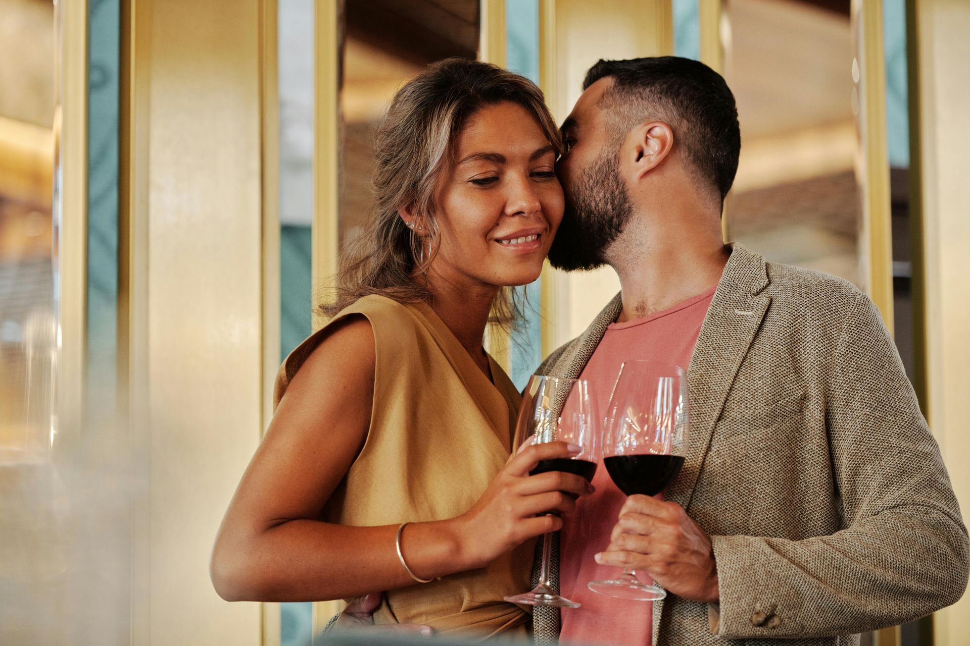 Man whispering into woman's ear, both holding wine glasses, in a chic setting.