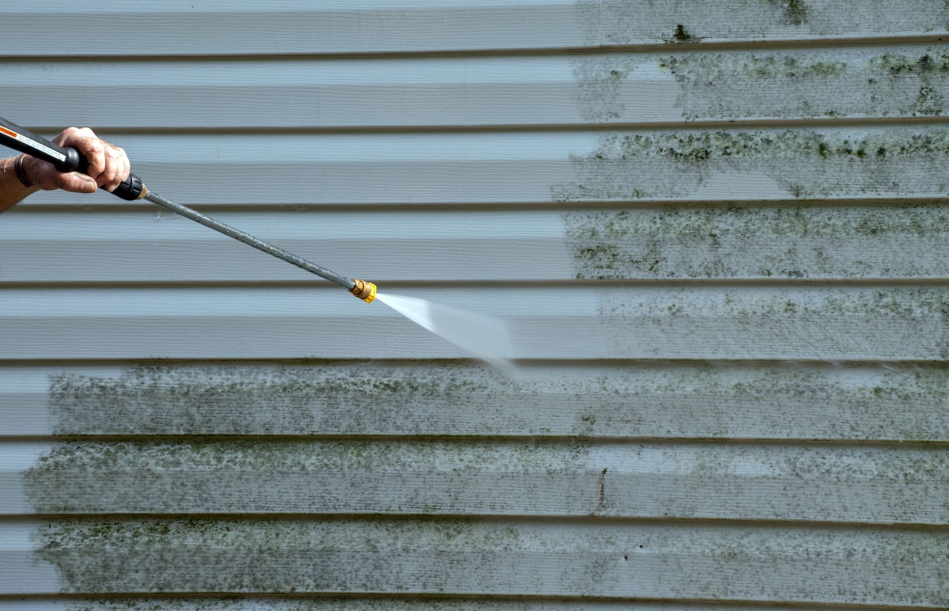 Person pressure-washing dirty gray vinyl siding with a spray wand. Person pressure-washing dirty gray vinyl siding with a spray wand.