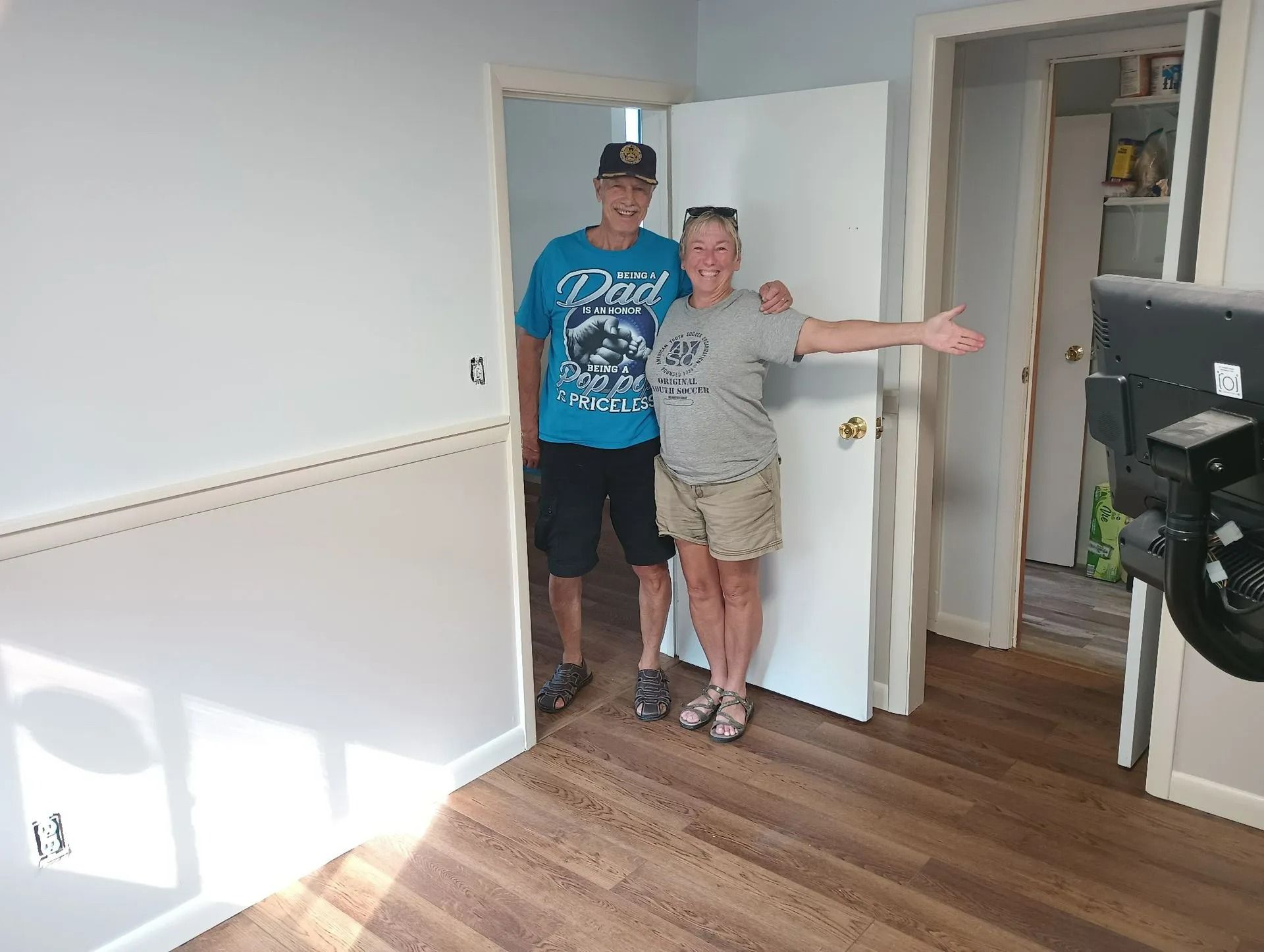 Two people standing in a bright room near a doorway, one smiling with arms outstretched