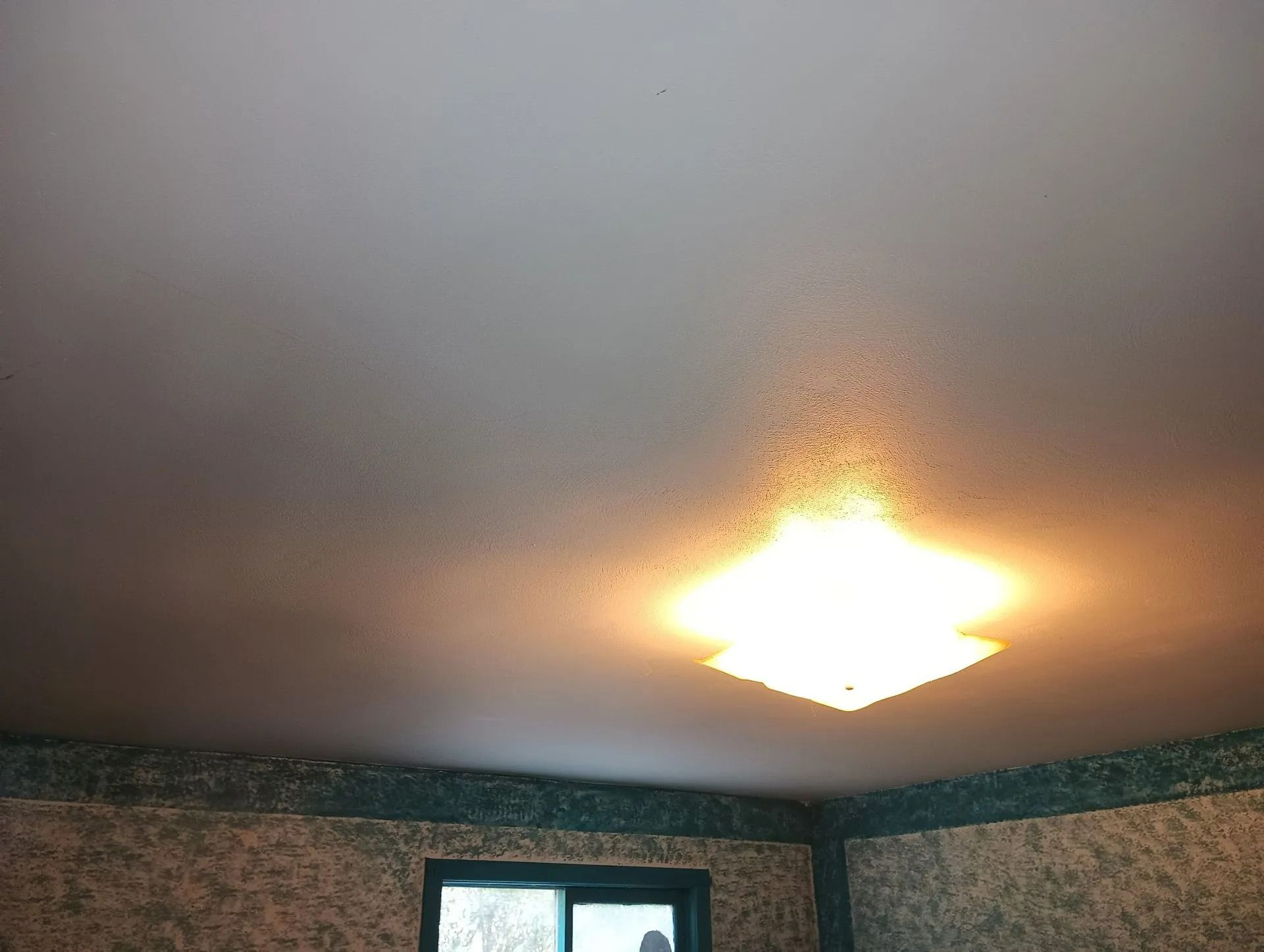 Ceiling with a bright flush-mount light over patterned wallpaper and a window opening