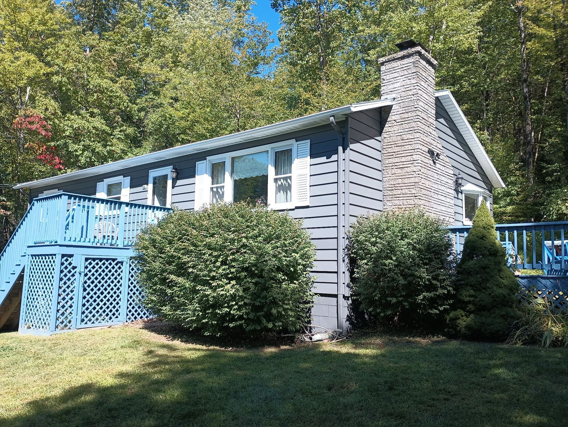 Small gray cottage with blue deck and white brick chimney, set among trees and shrubs.