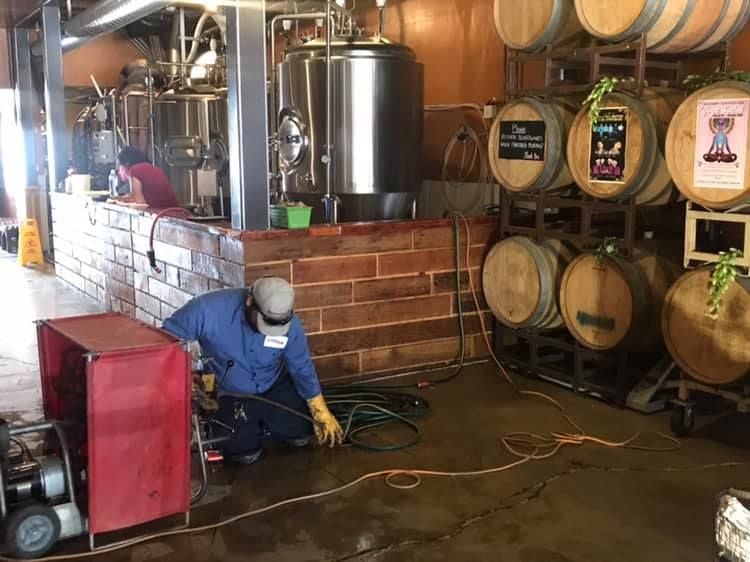 Worker cleans brewery floor beside beer barrels and stainless tanks