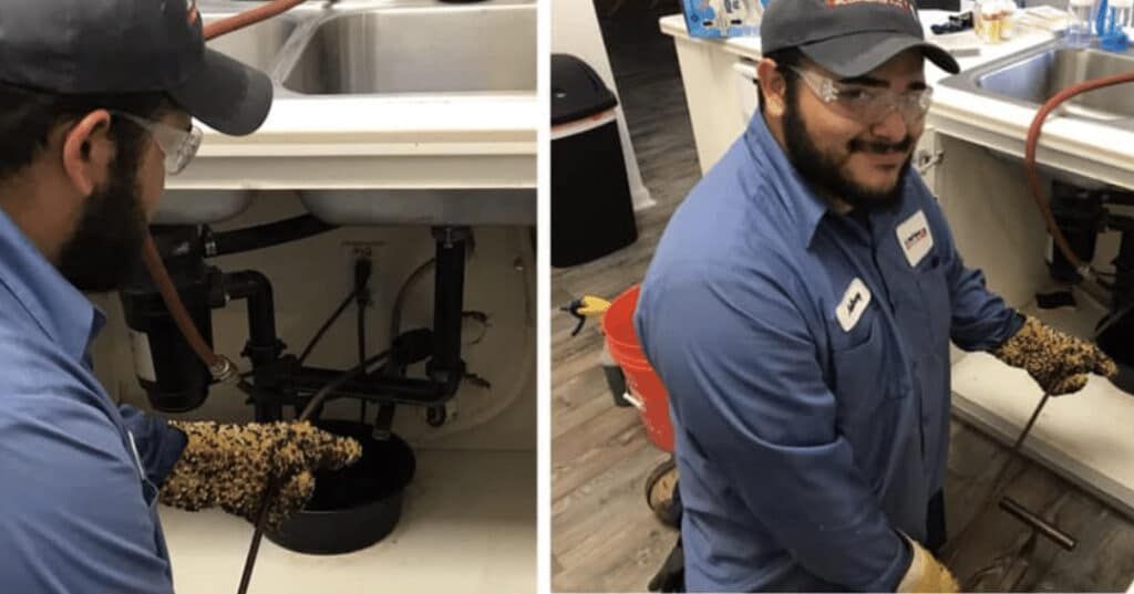 Technician in blue shirt and cap working under a sink, smiling at the camera, in a workshop.