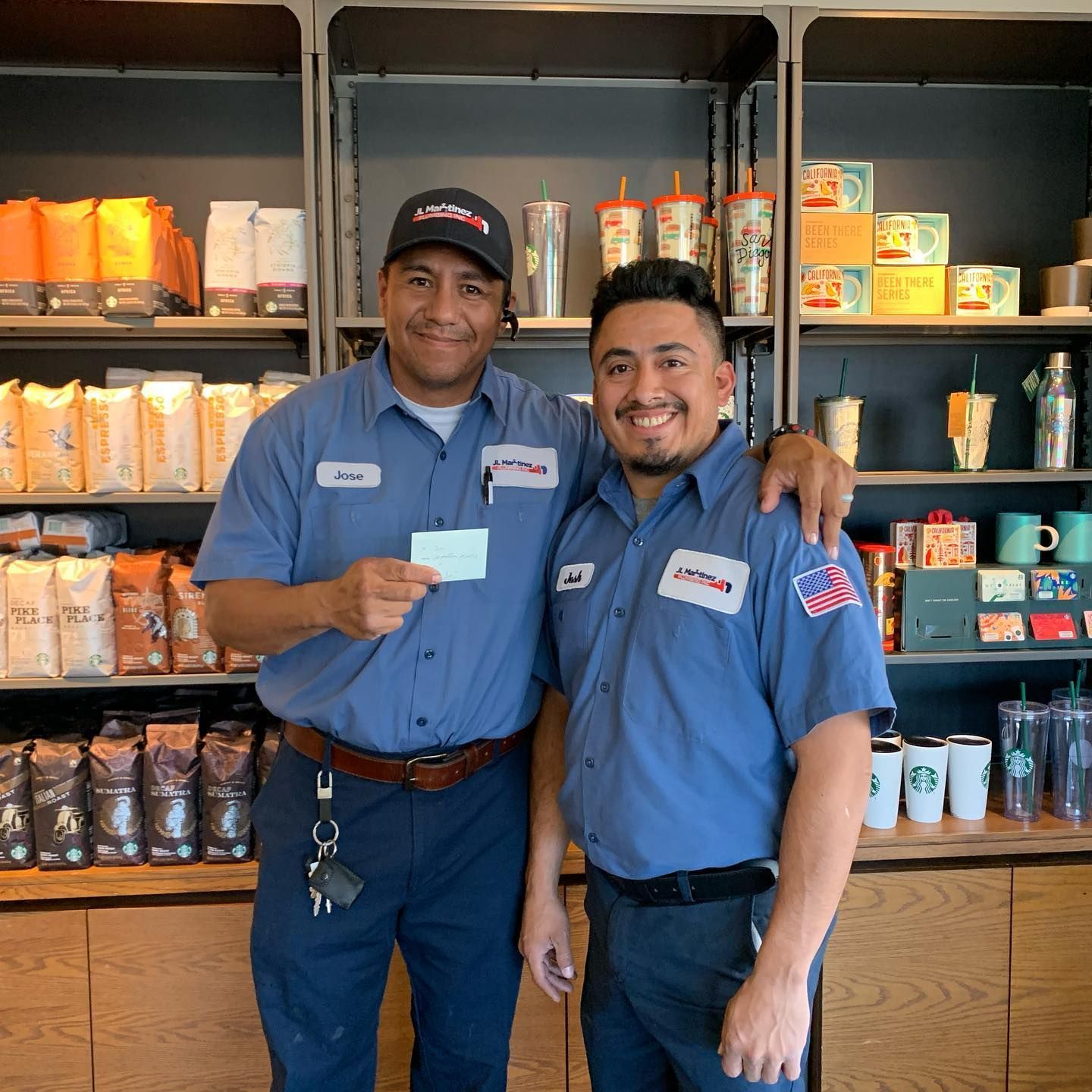 Two store employees in blue uniforms smiling behind a café counter with shelves of drinks and snacks.
