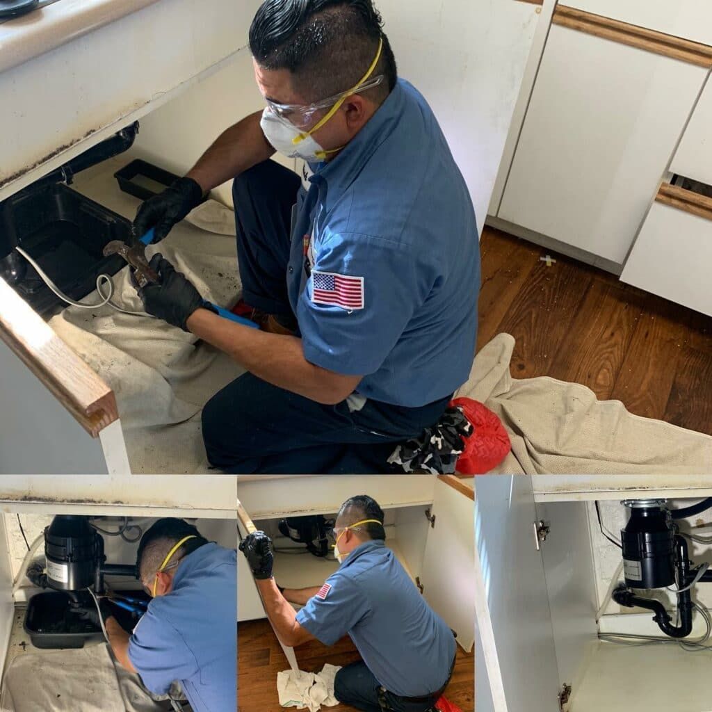 Plumber repairing a kitchen sink cabinet, wearing gloves, mask, and safety glasses.