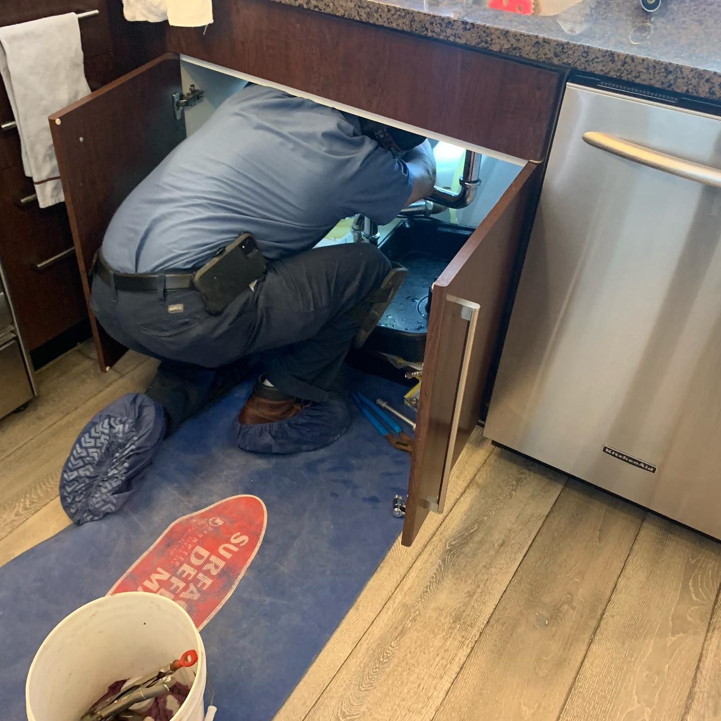 Plumber working under a kitchen sink with cabinet doors open, on a blue protective mat.