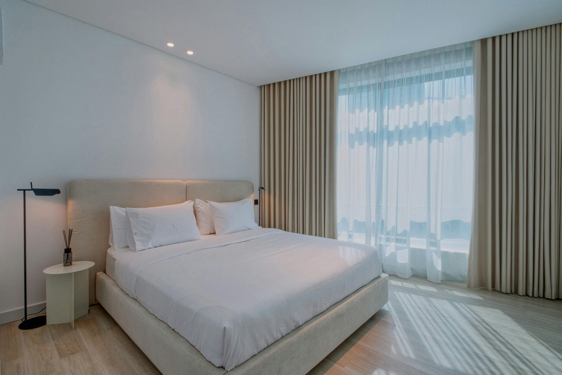 A modern bedroom featuring a large white bed, beige curtains over a window, and a minimalist floor lamp on a side table.