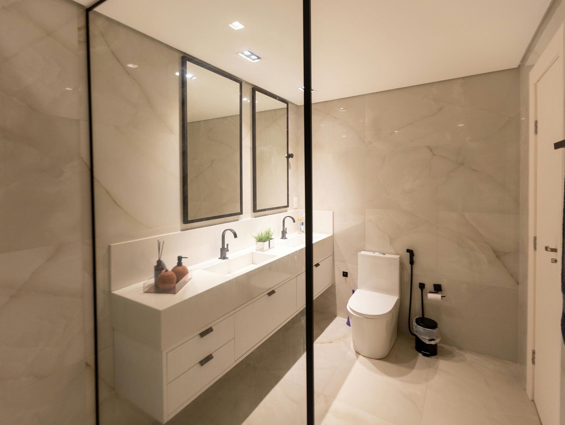 Modern bathroom with white countertops, sinks, mirrors, and toilet, black accents, and marble-look tile walls.