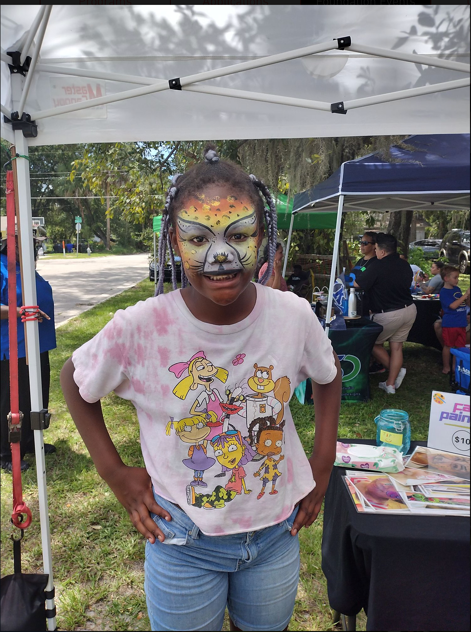 A young girl with her face painted is standing in front of a tent.