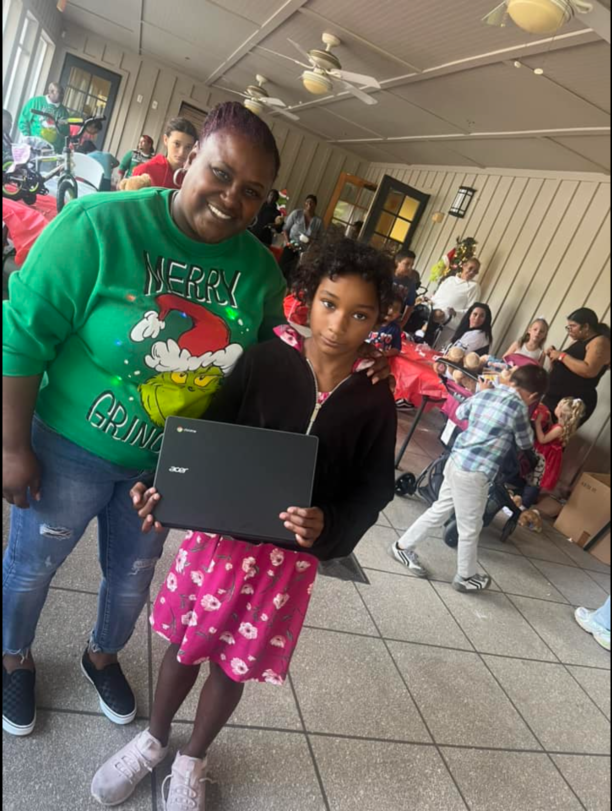 A woman in a green grinch sweater is standing next to a little girl holding a laptop.