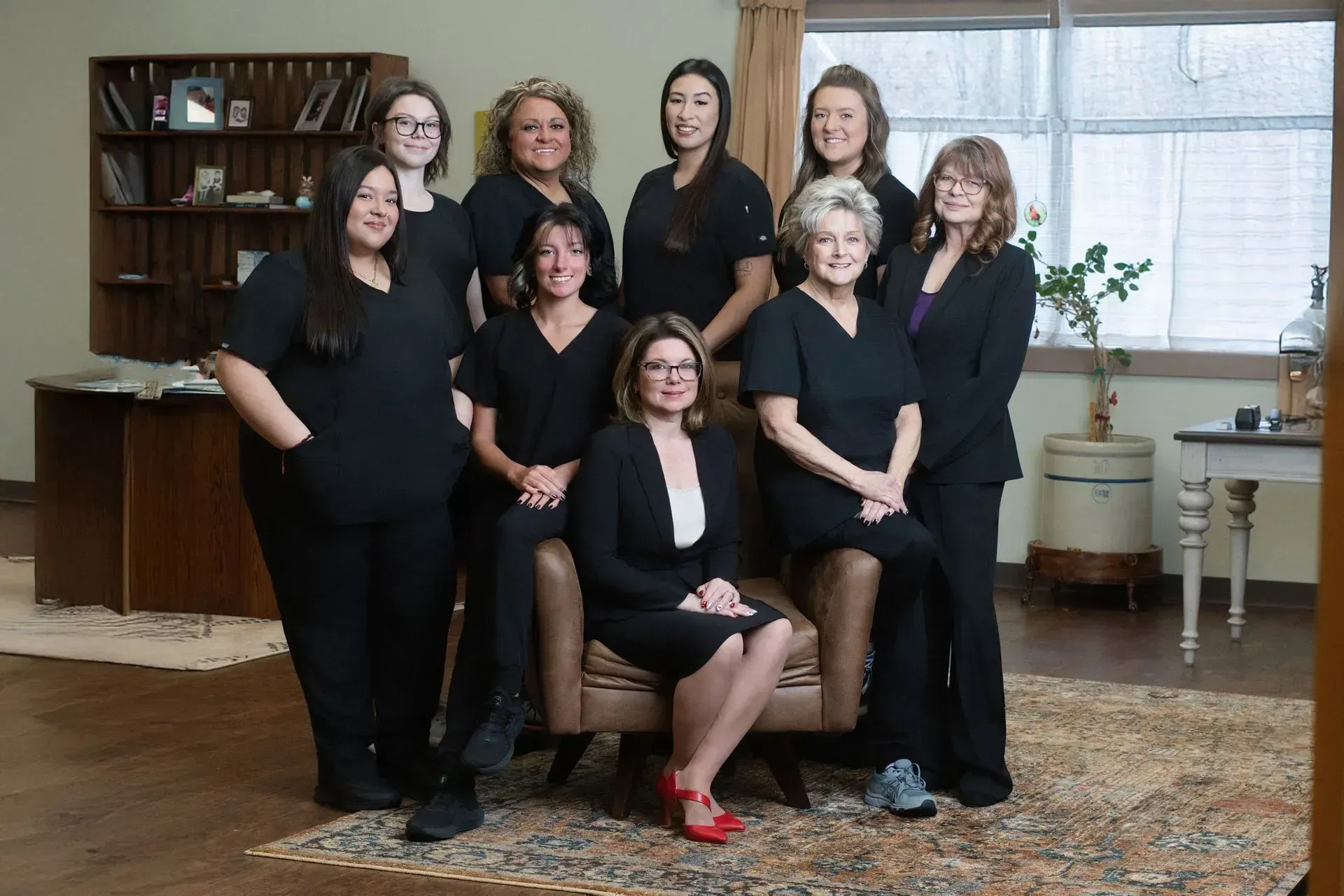 Group of eight women posing in a living room, dressed in black, with one seated in red shoes.