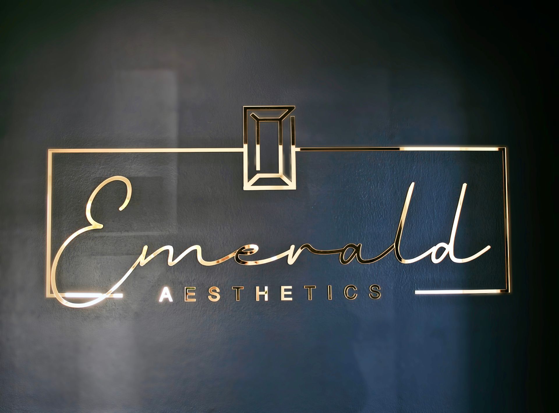 Gold “Emerald Aesthetics” sign on a dark wall with a minimalist square logo.