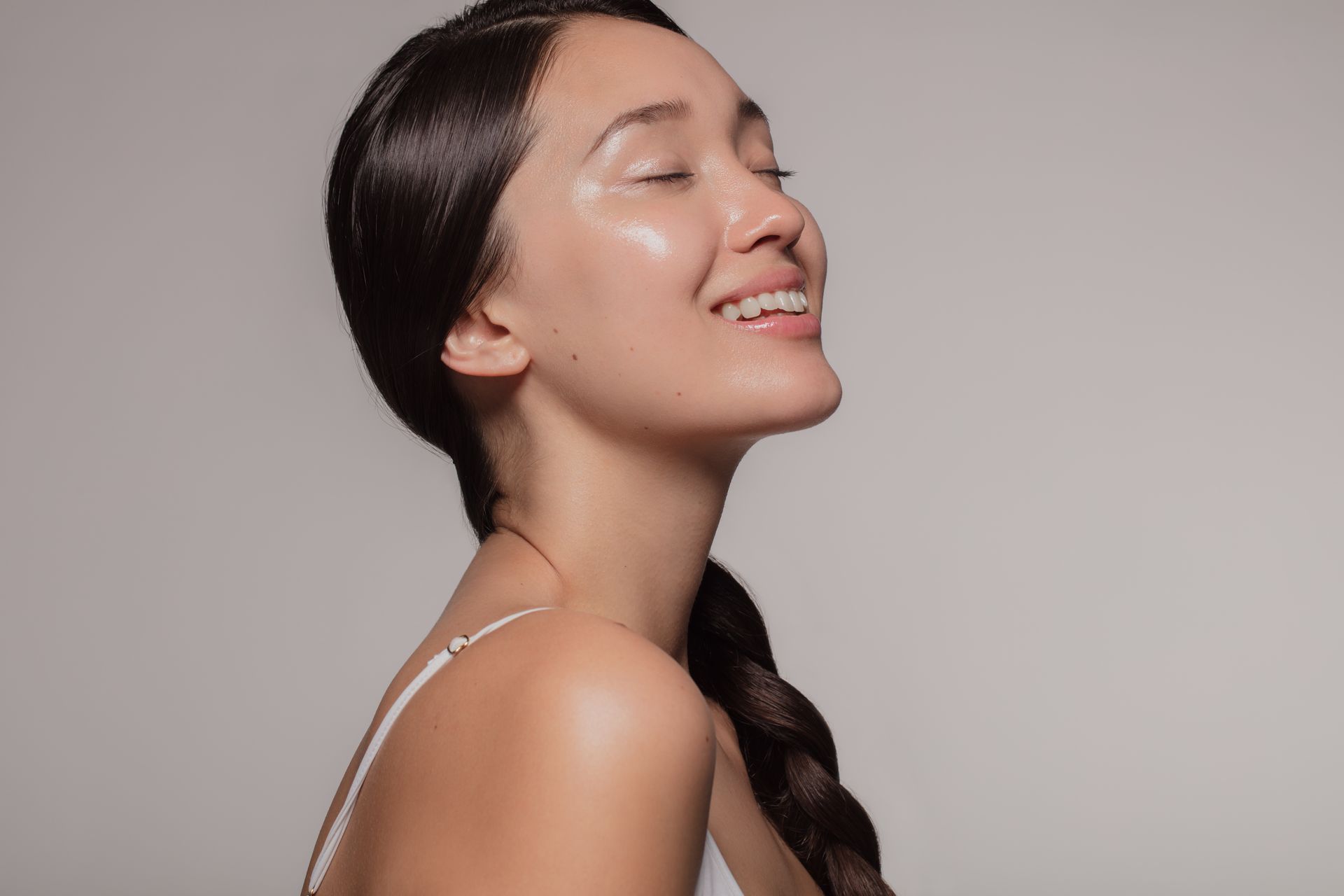 Woman with shiny skin, smiling, eyes closed, hair in a side braid, wearing a white tank top.