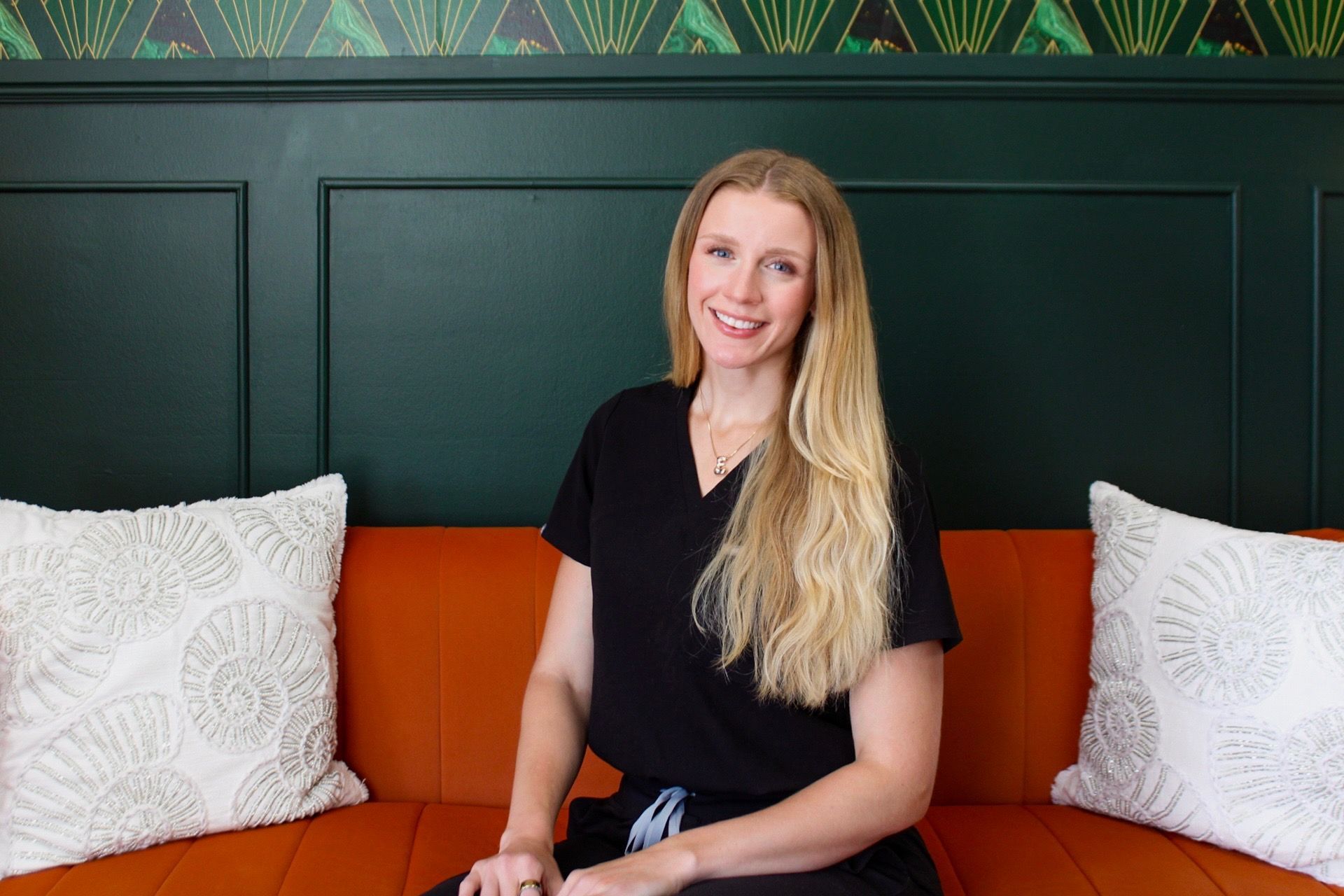 Woman seated on an orange sofa between white pillows, smiling in front of a dark green wall.