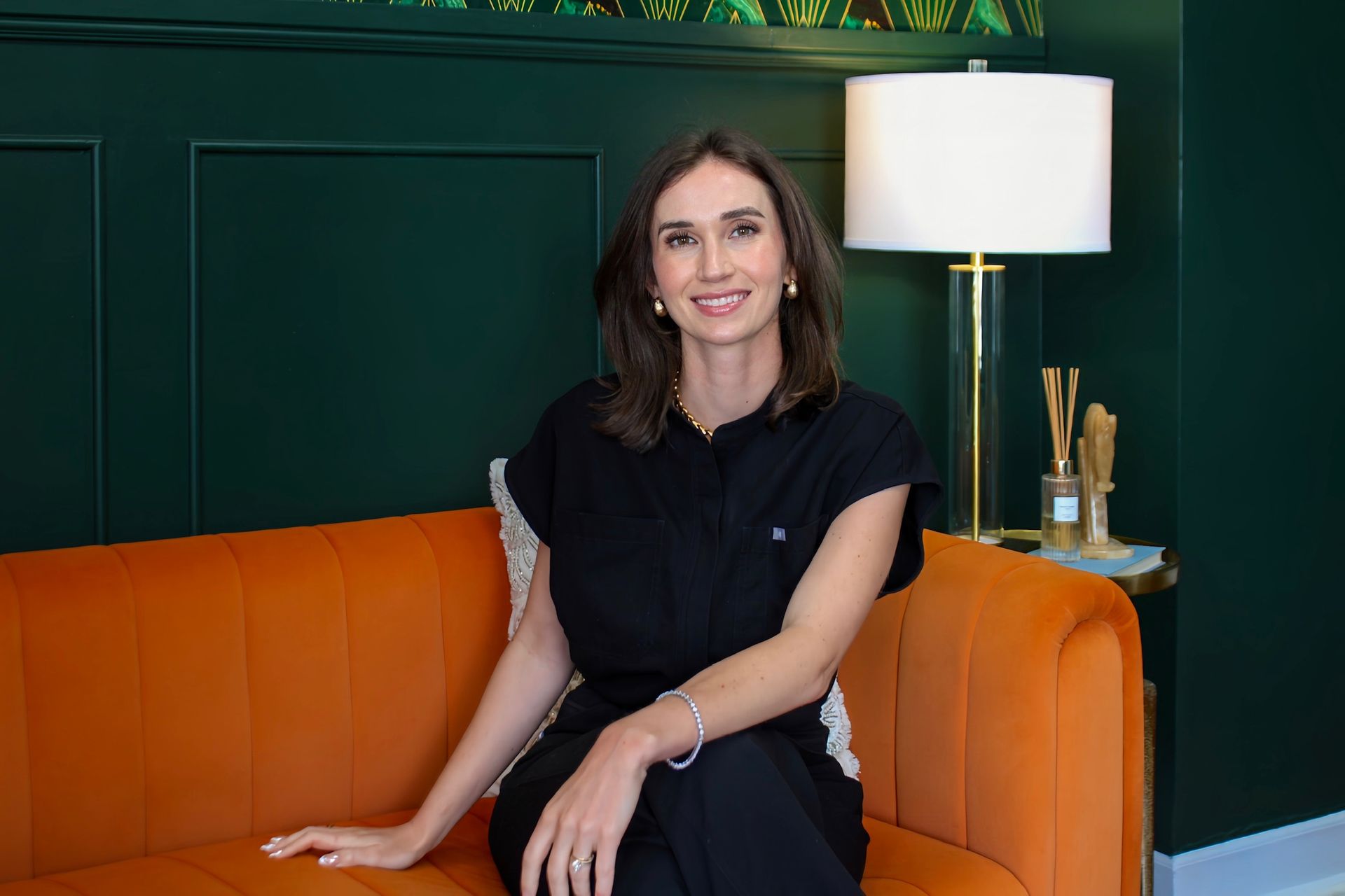 Woman seated on an orange sofa in a dark green room beside a lit floor lamp