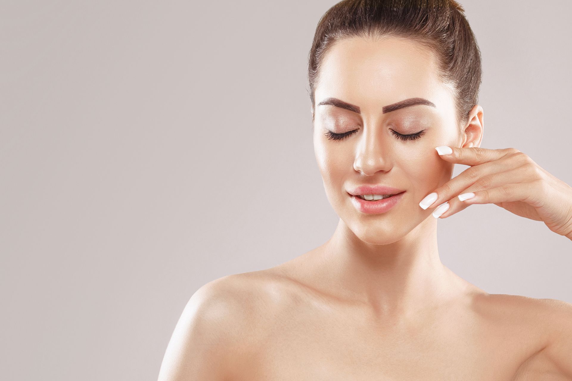 Woman with eyes closed applying cream to her cheek against a plain beige background