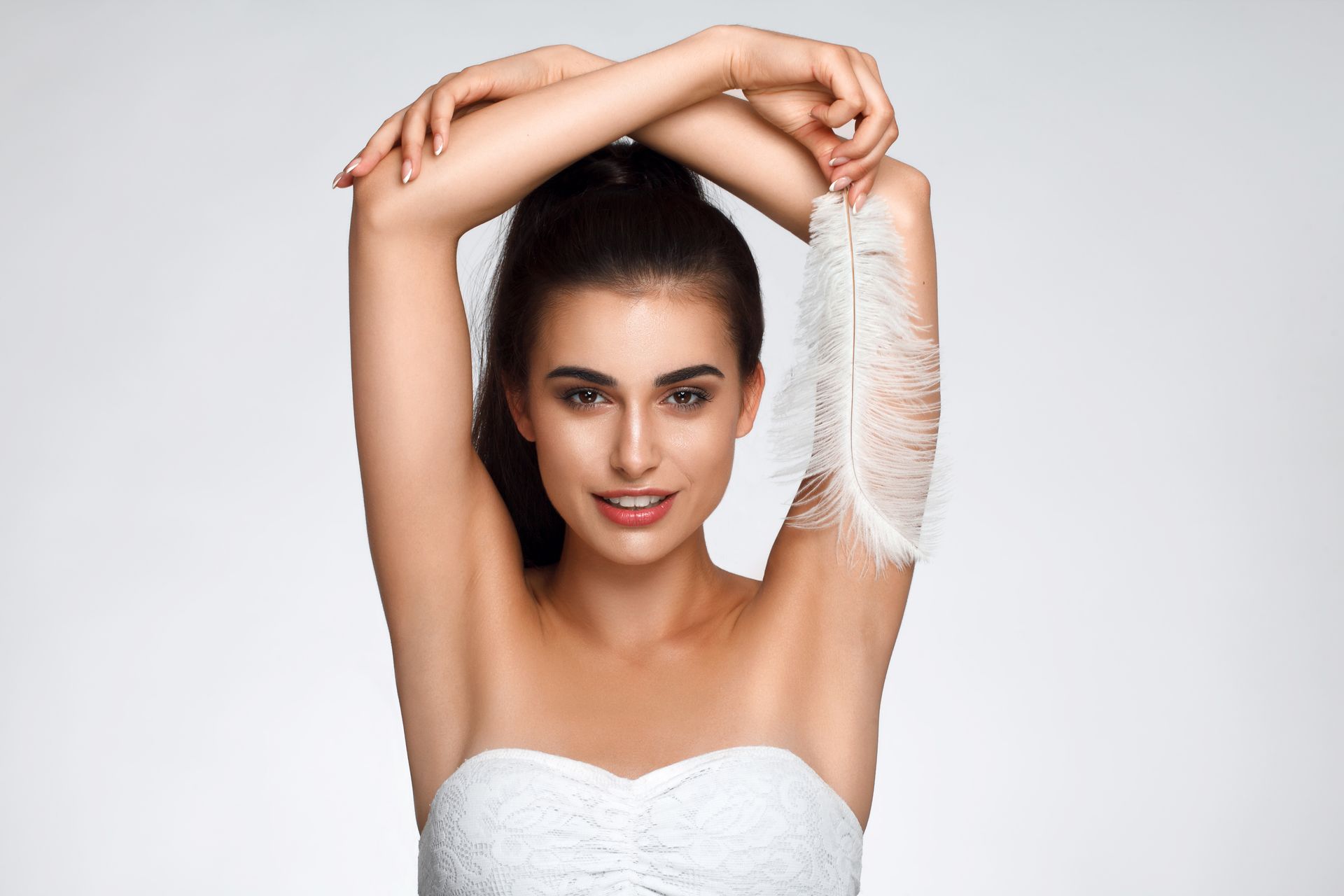 Woman holding a feather to her underarm, smiling, in front of a light background.