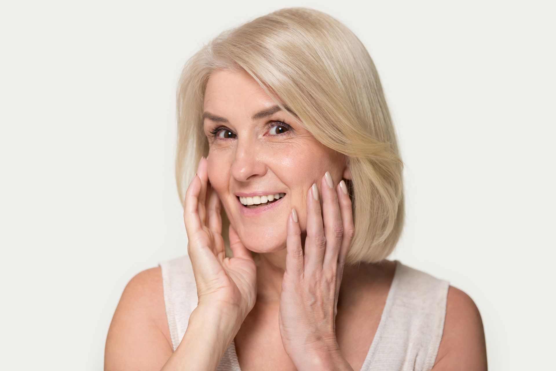 Woman with blonde hair smiling, touching her face, against a light background.
