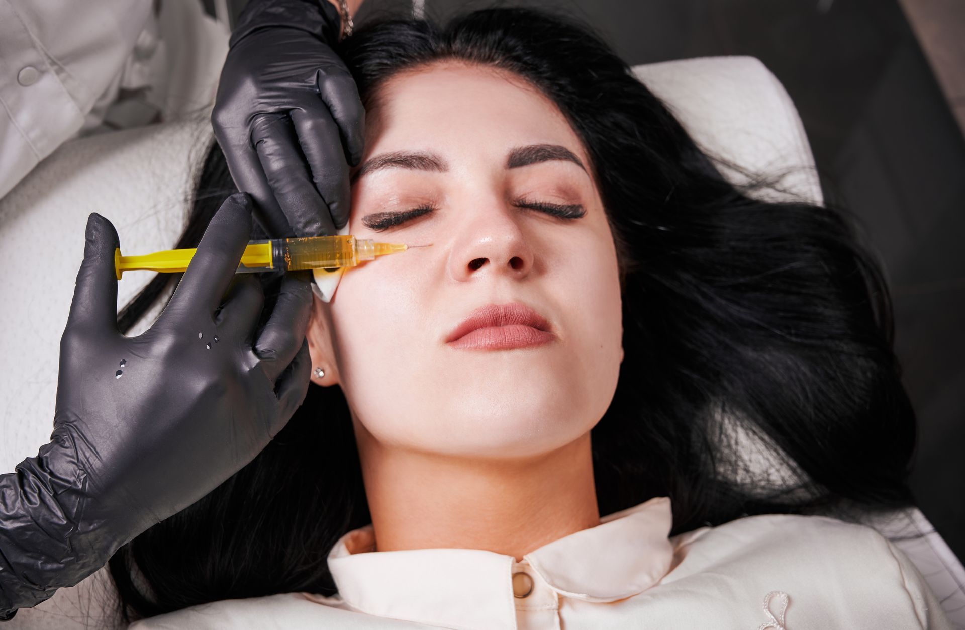 Gloved hands inject facial filler into a woman lying on a treatment bed