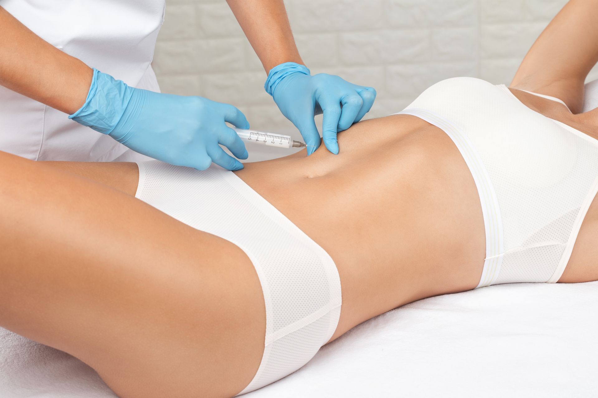 Medical professional injecting medication into a person's abdomen, wearing gloves and white uniform, setting is a clinic.
