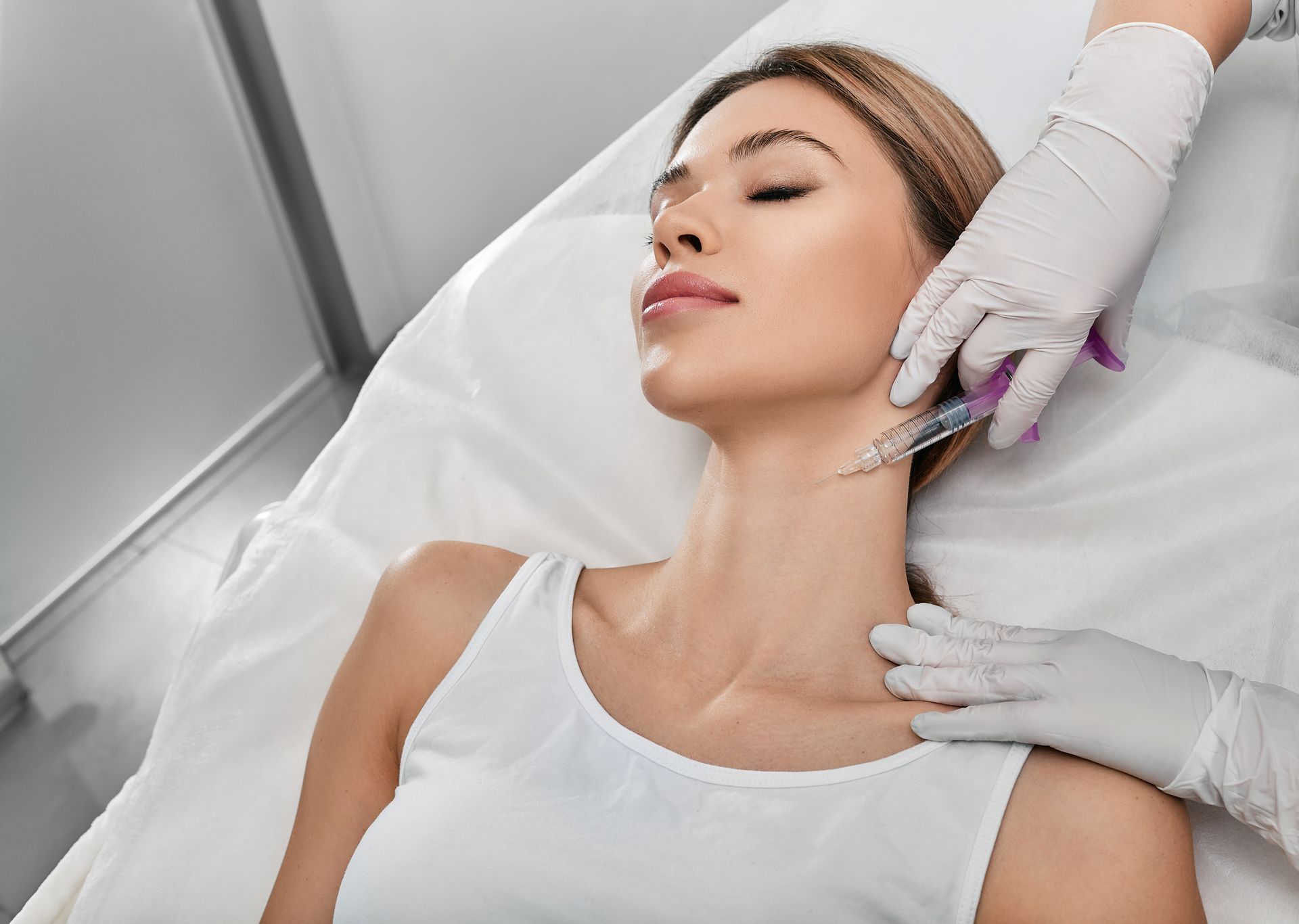 Woman receiving neck injections from a gloved hand, lying on a white bed in a clinic.