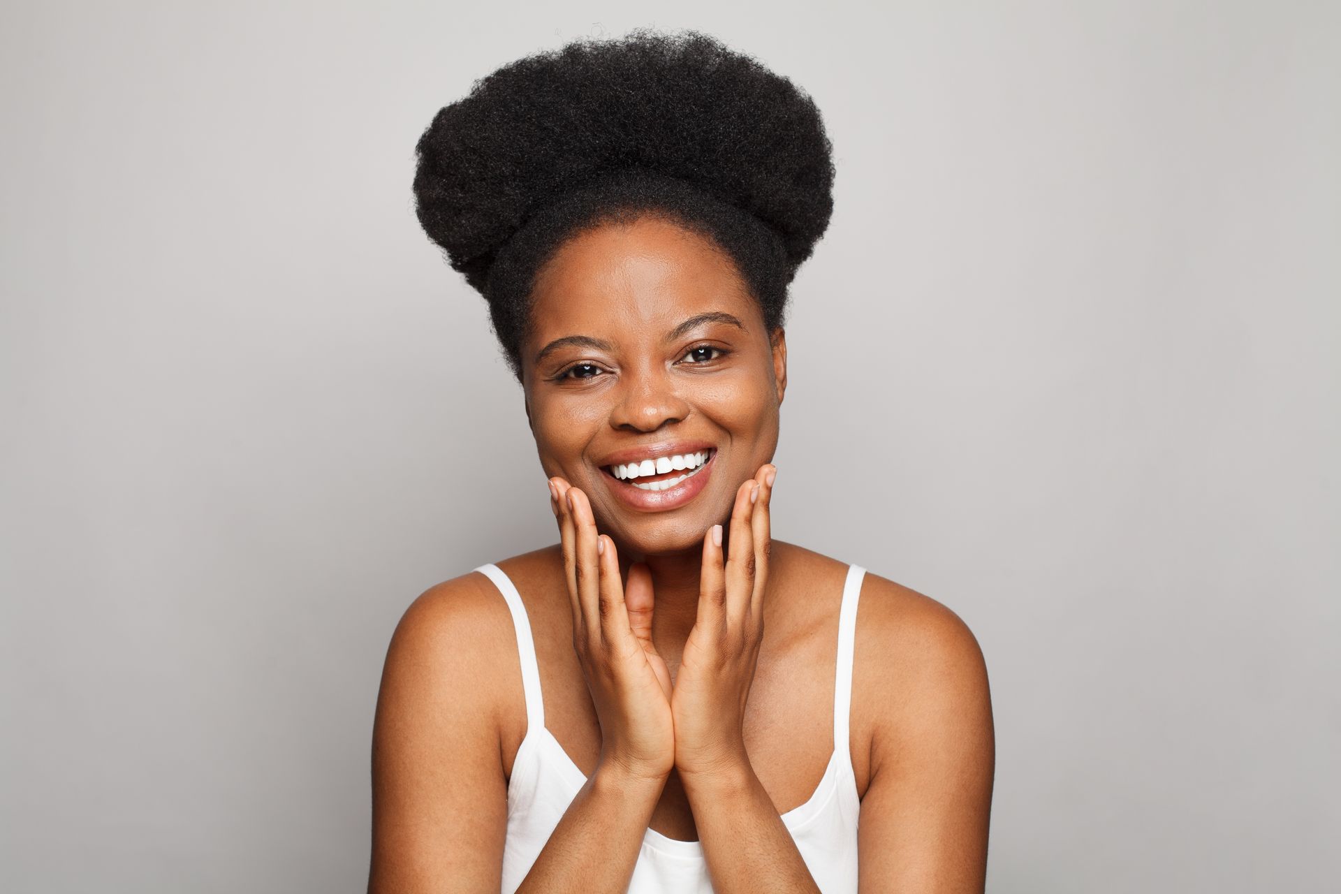 Smiling person in a white tank top, hands on cheeks, against a gray background